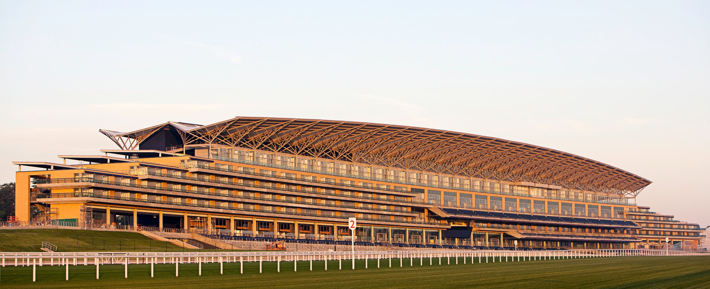 Ascot's grandstand in early morning light, its curved roof above the empty straight and white running rail.
