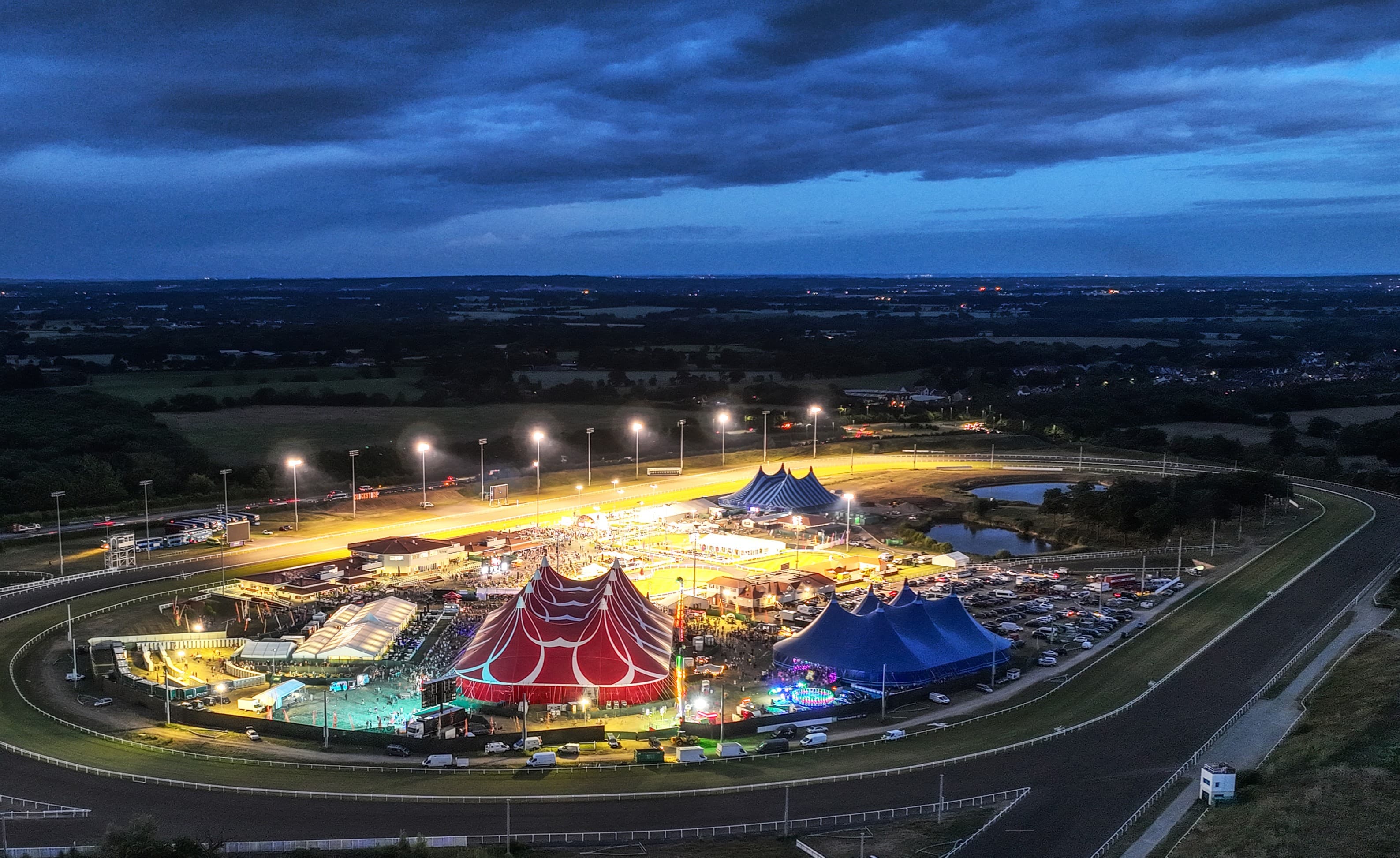 Aerial view at dusk of the floodlit all-weather oval at Chelmsford City Racecourse, with marquees and a funfair filling the infield.