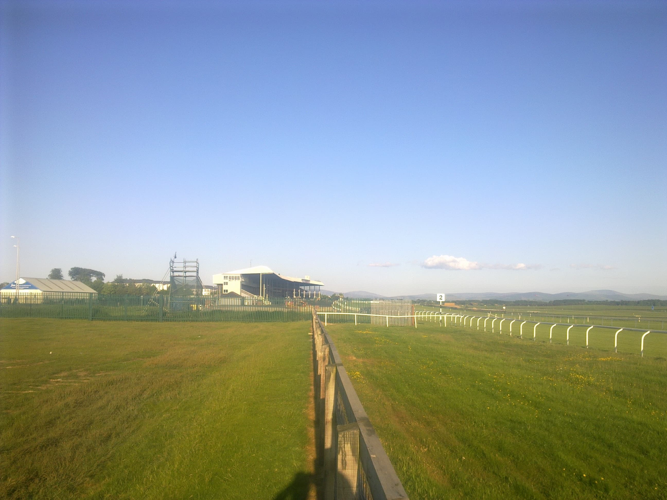 The Curragh racecourse on a clear summer evening, the running rail leading towards the grandstand with the Kildare plain beyond.