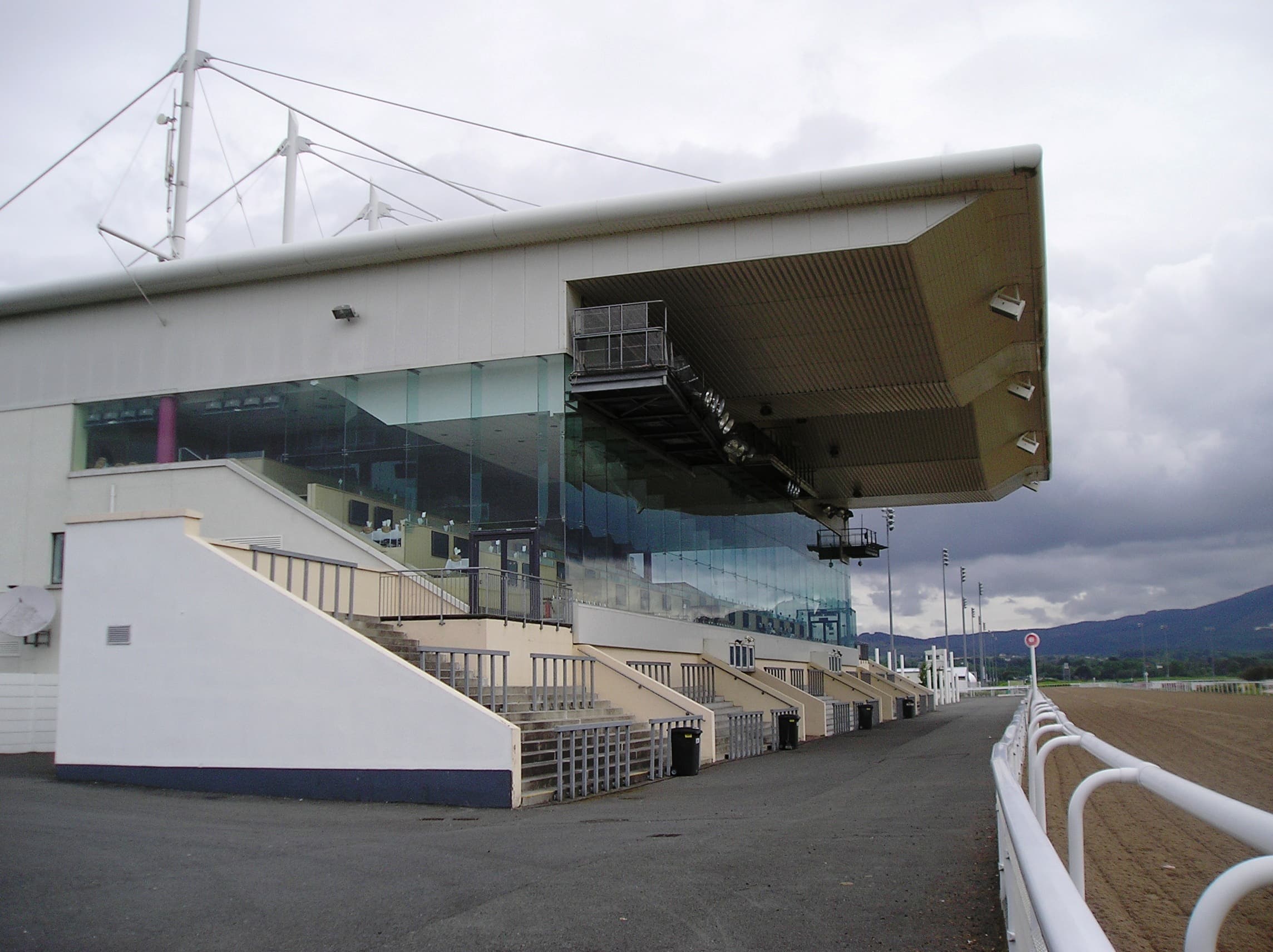 The cantilevered glass grandstand at Dundalk Stadium seen along the rail, the sand track running away in front of it.