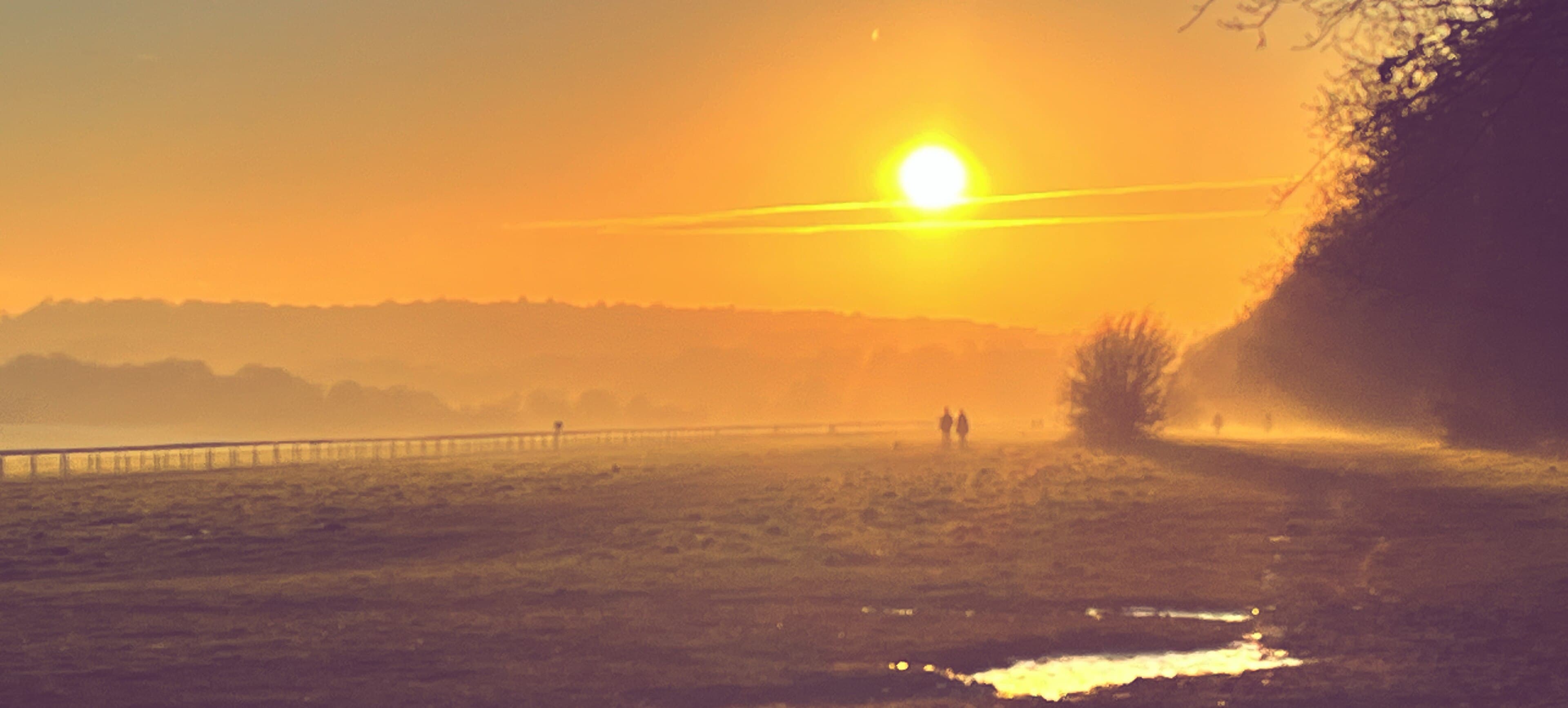 Sunrise through mist over the track at Epsom Downs, with two walkers in silhouette on the far side.