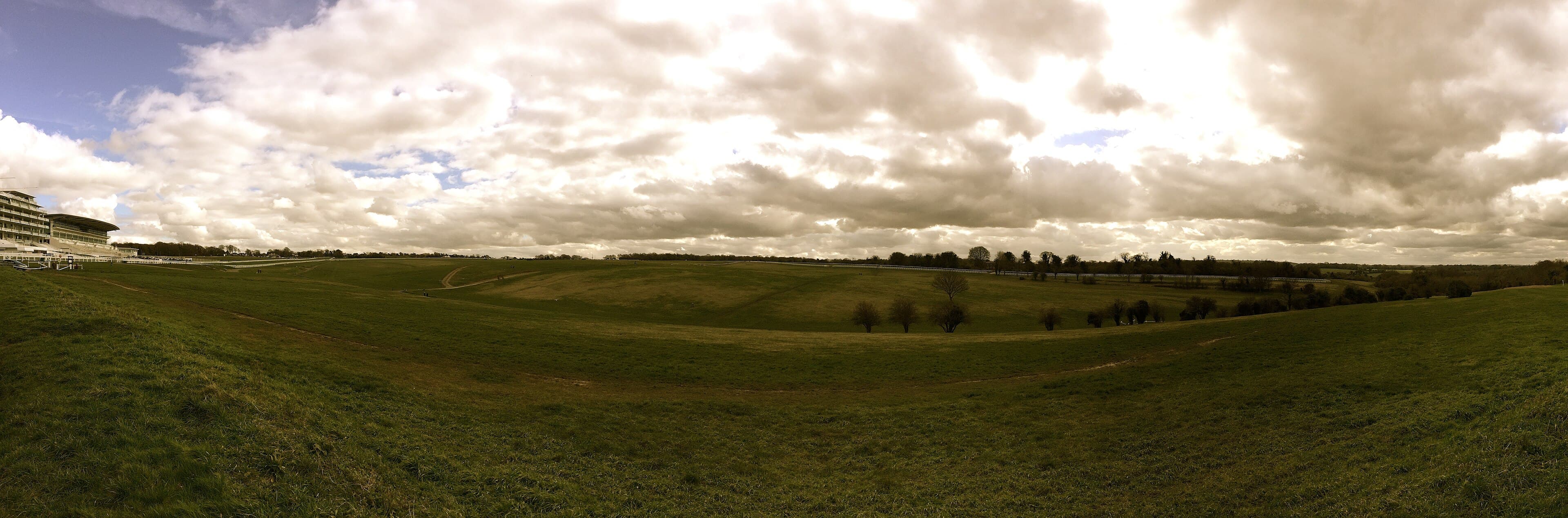 A wide view over Epsom Downs with the grandstand on the skyline to the left and broken cloud above the course.