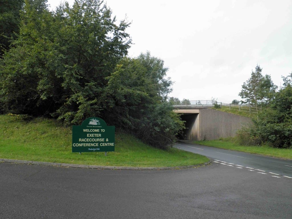 The green Welcome to Exeter Racecourse and Conference Centre sign on the grass verge at the access road turning, with the A38 underpass beyond.