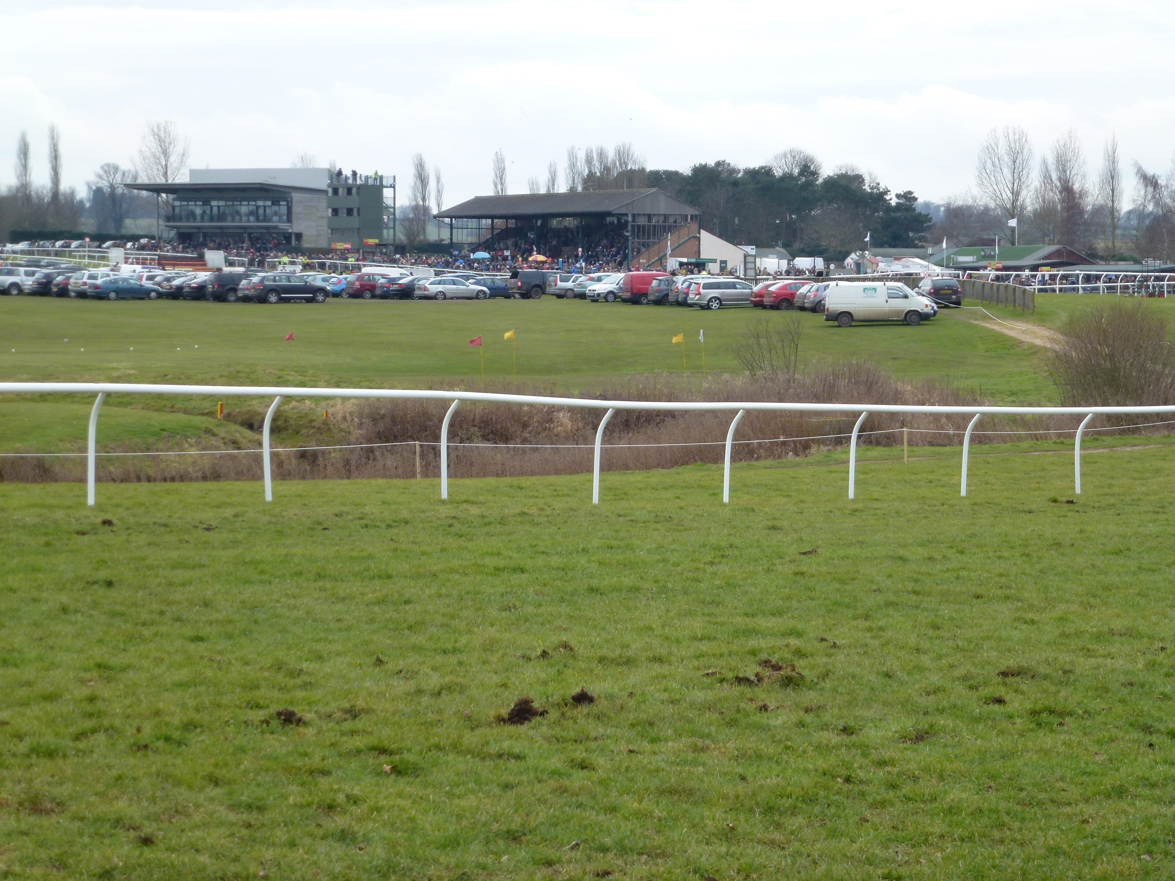 A wide view across the course at Fakenham to the modern grandstand and the older covered stand, with cars parked along the rail.