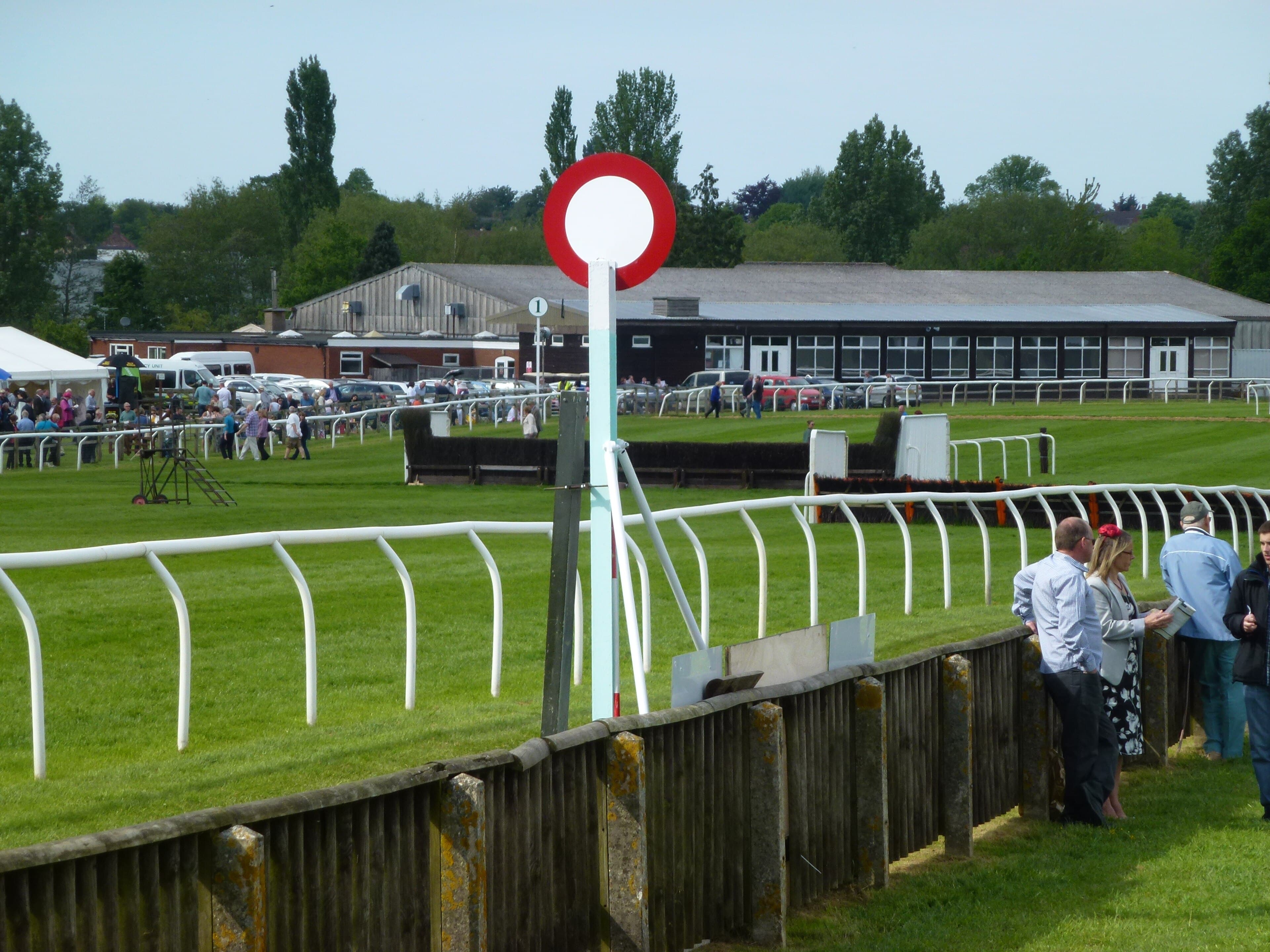 The red and white winning post at Fakenham, with the running rail, a hurdle and the low course buildings behind.