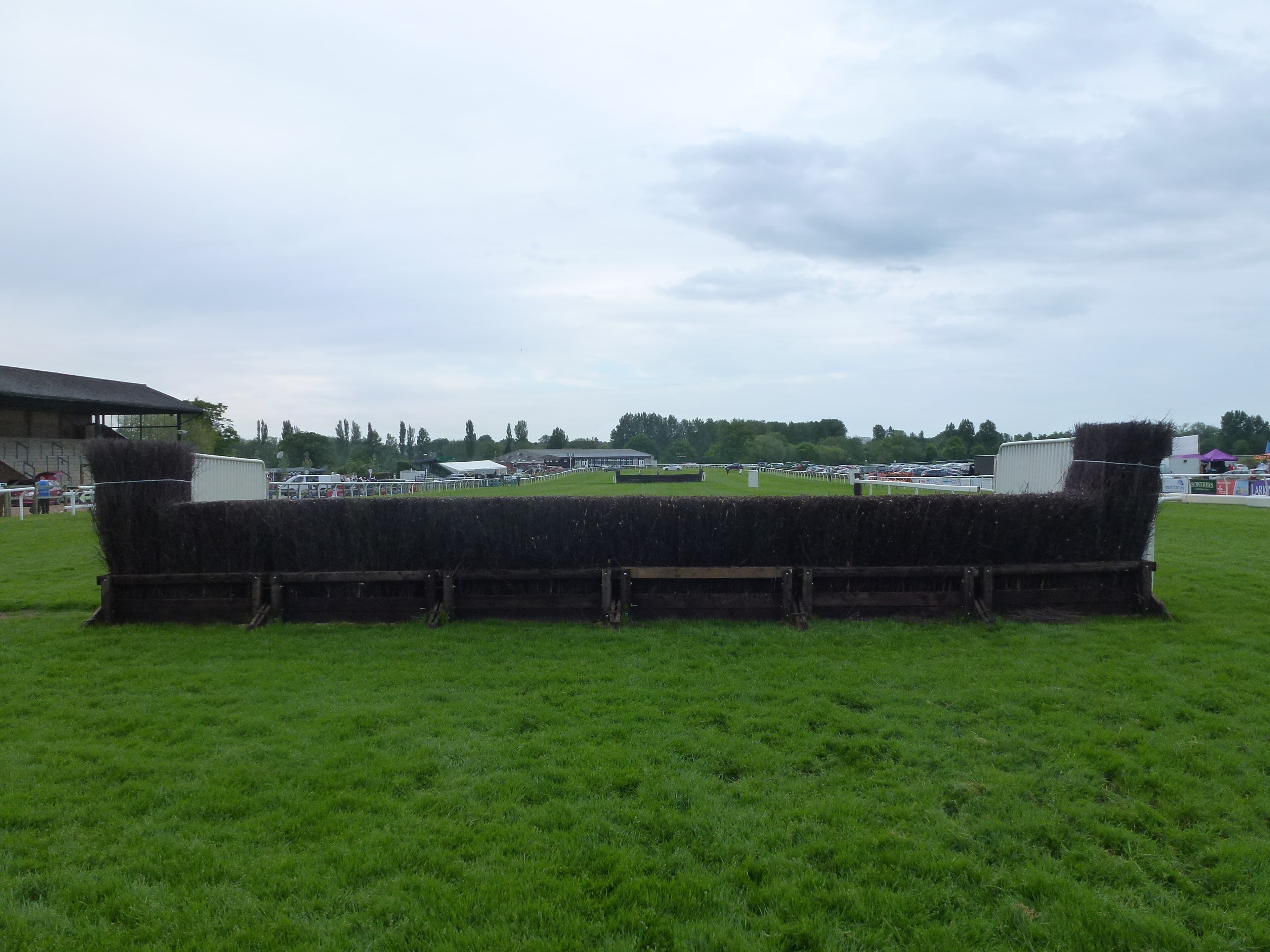 A birch steeplechase fence at Fakenham photographed head on across empty turf, with the course buildings small in the distance.