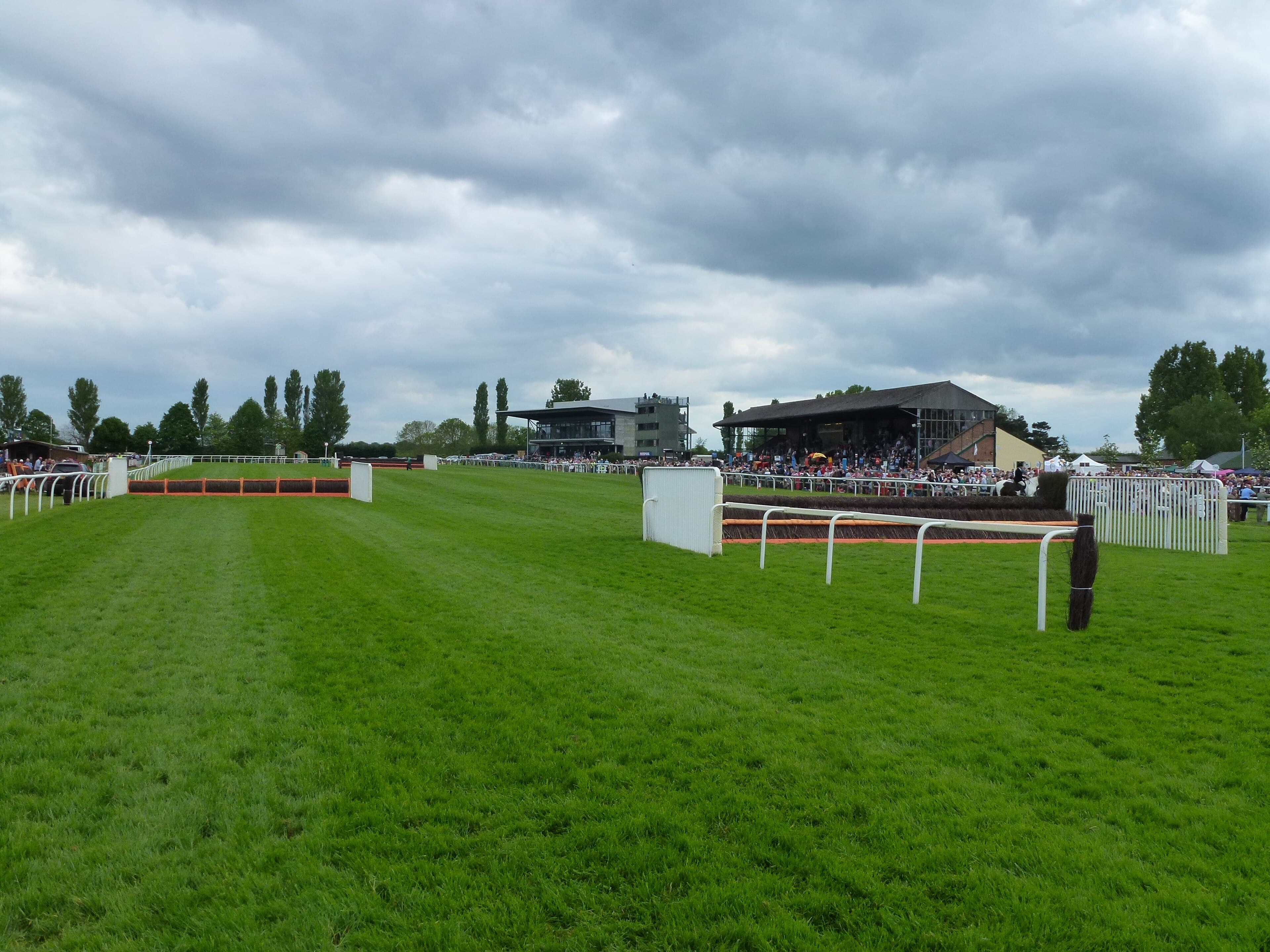 The finishing straight at Fakenham with two hurdles in the foreground and both grandstands and a raceday crowd behind.