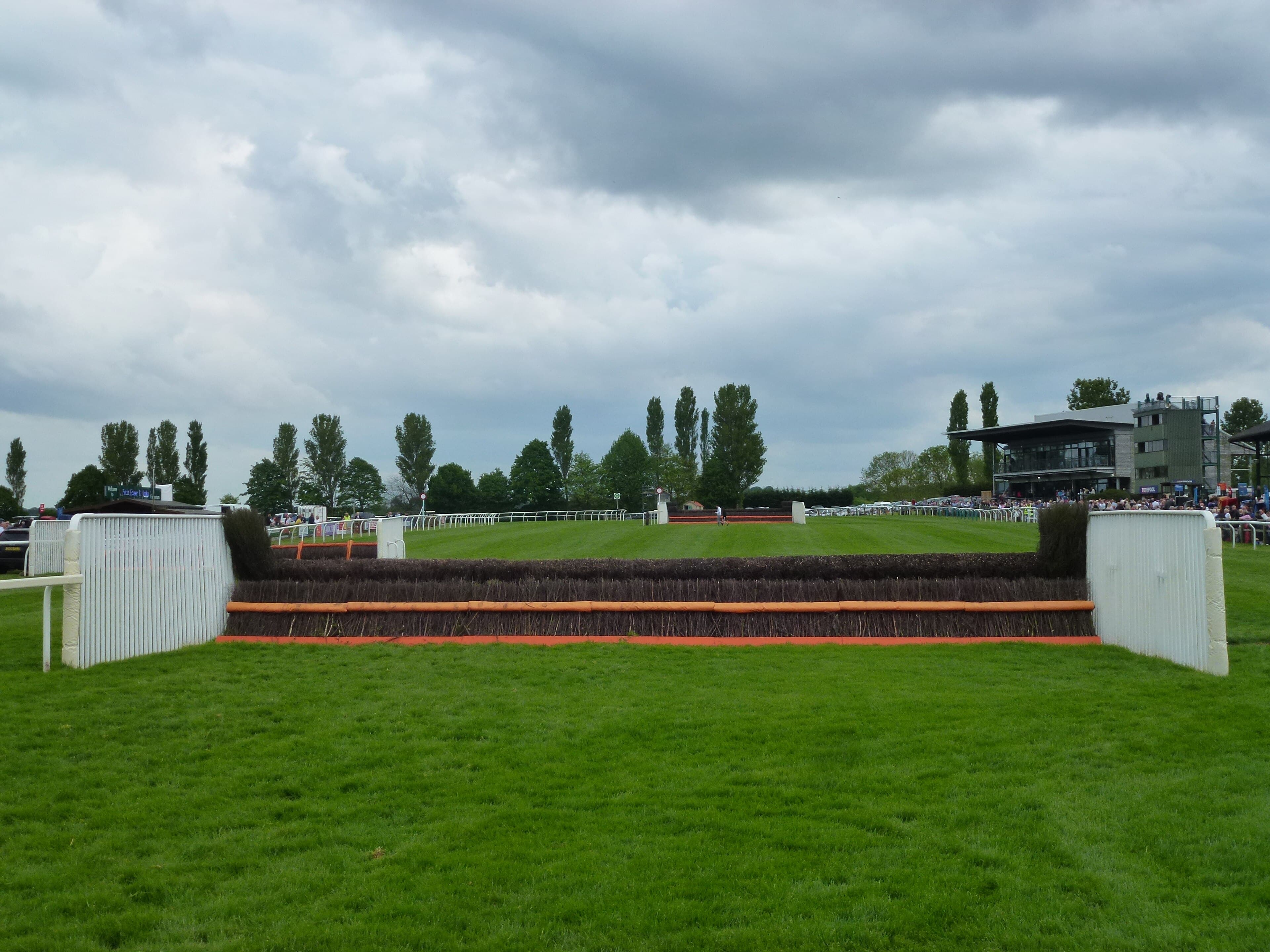 A birch chase fence at Fakenham seen straight on, with the glass-fronted grandstand and a crowd behind it across the course.