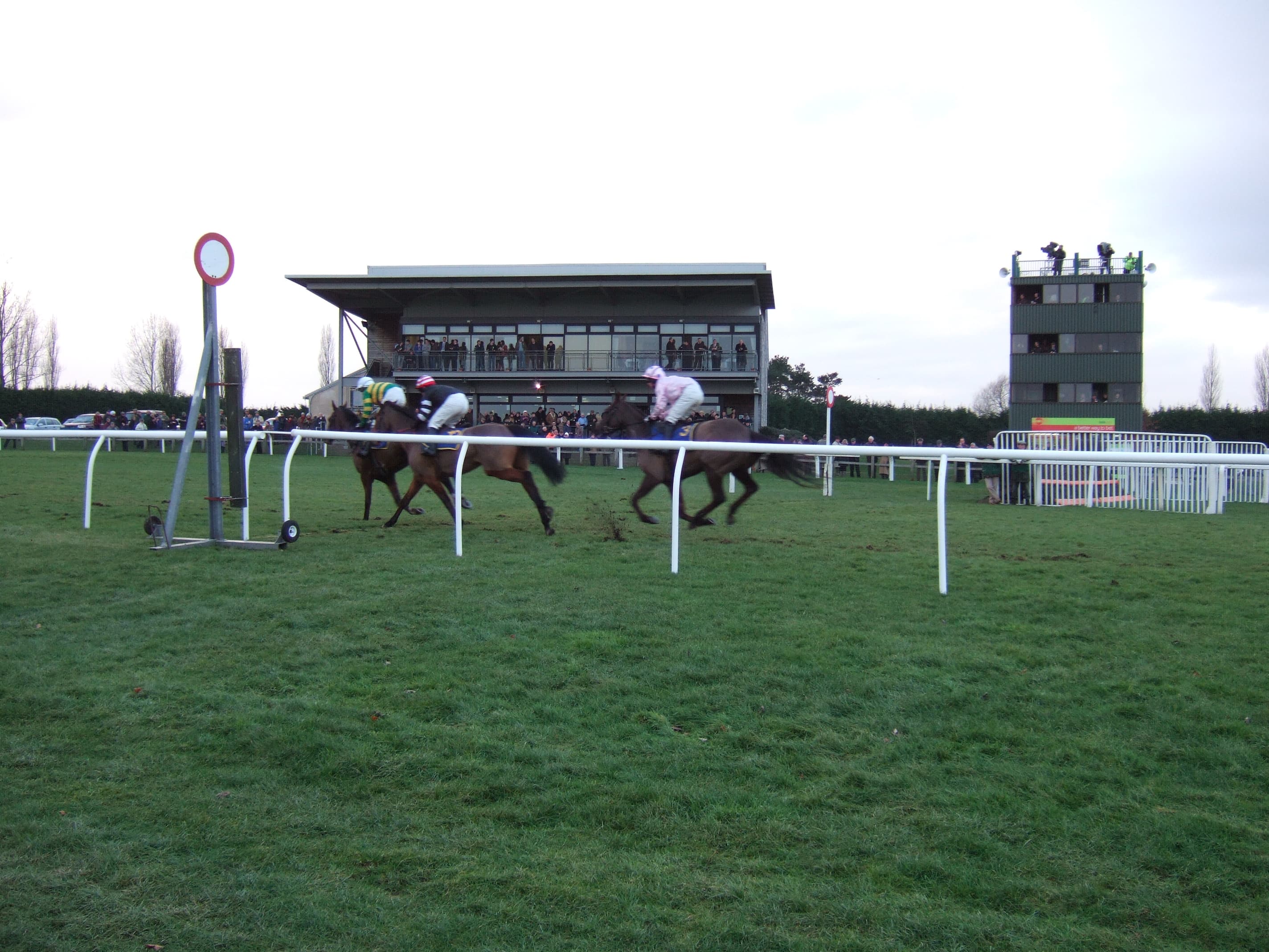 Three runners passing the winning post at Fakenham with the glass-fronted grandstand and judge's box behind.