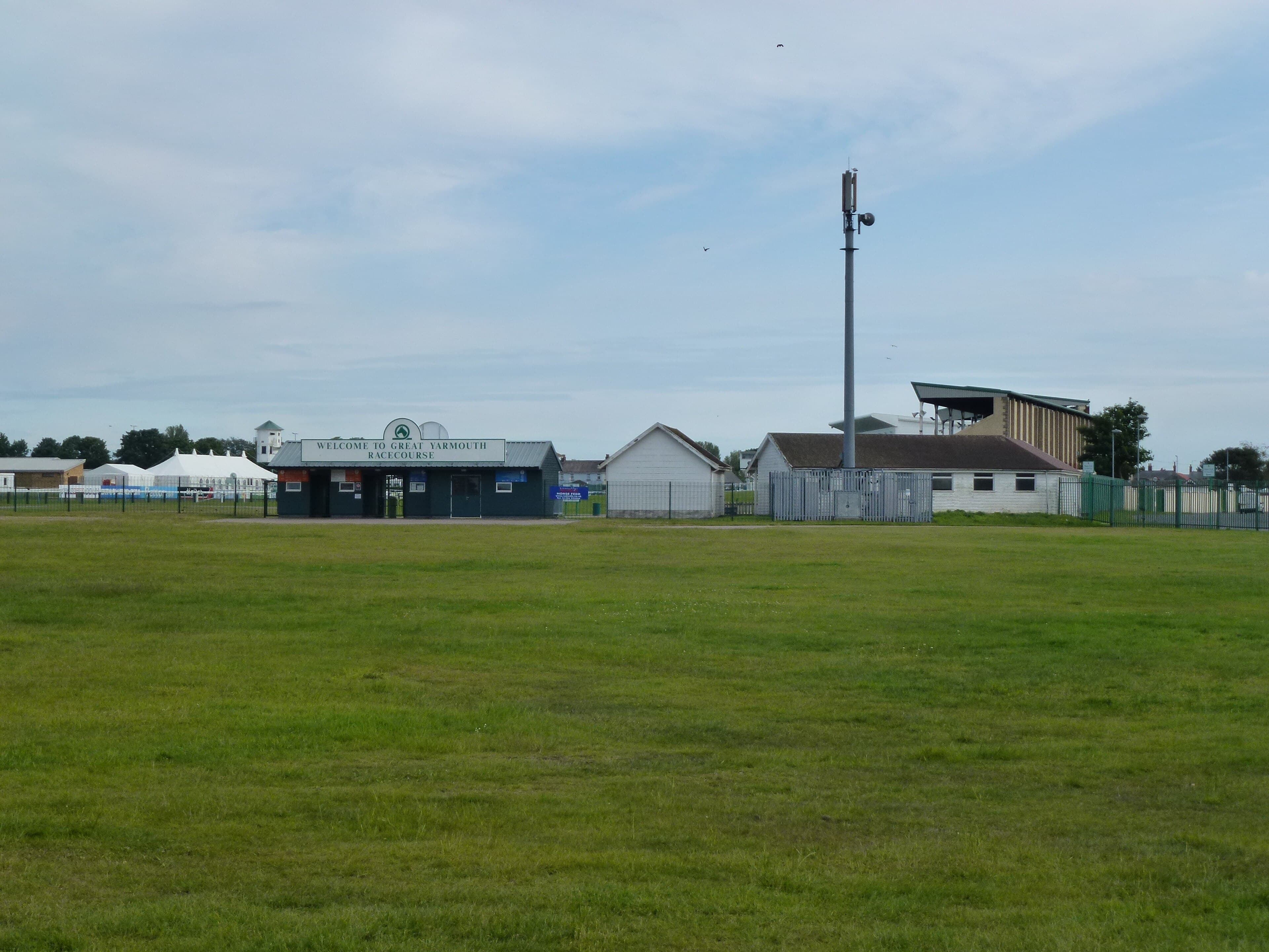 The green entrance building at Great Yarmouth racecourse with turnstiles under a Welcome to Great Yarmouth Racecourse sign, the stand roof and a mast behind