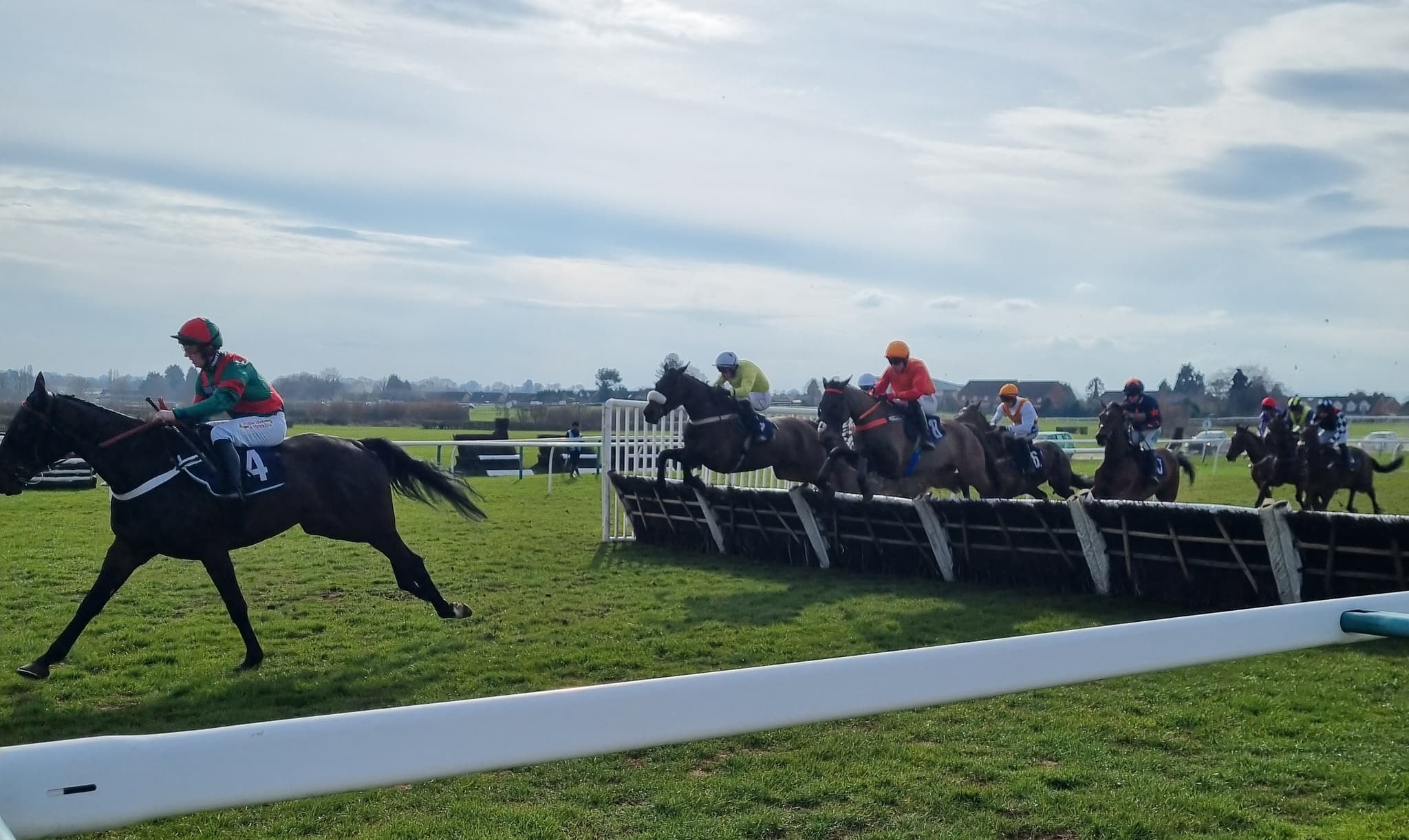 A big field of hurdlers jump a flight together at Hereford while the long-time leader gallops clear in front, the grandstand and open Herefordshire countryside beyond the rail.