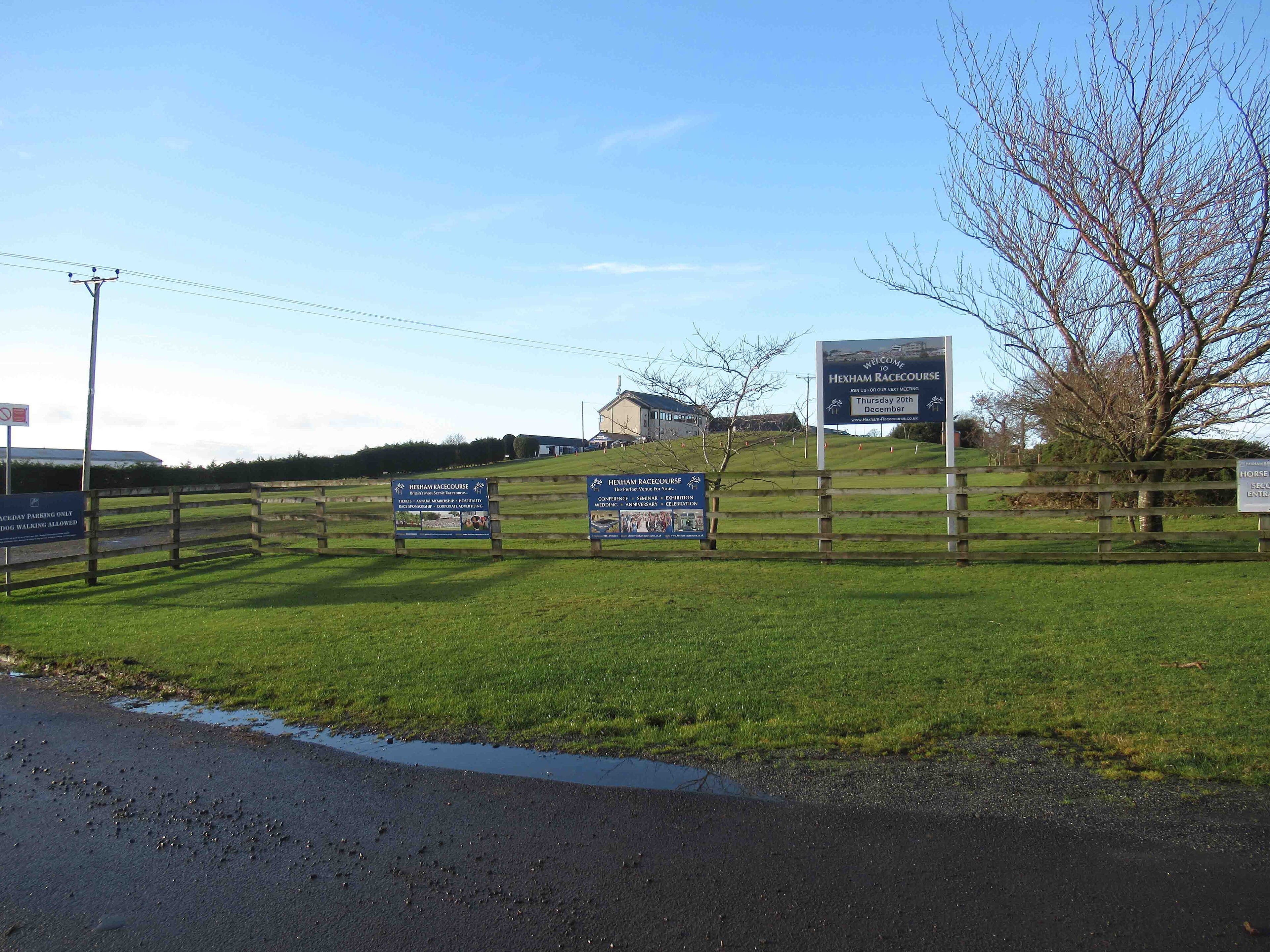 The roadside welcome sign at the entrance to Hexham Racecourse, with the main stand on the hill behind and racecourse advertising boards along a post-and-rail fence.