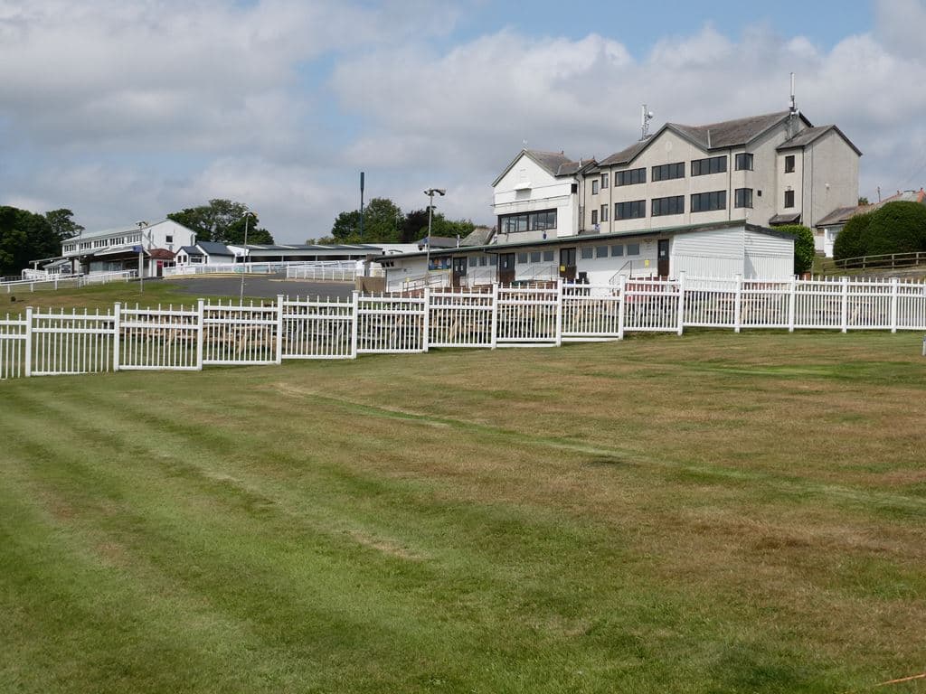 The main grandstand and weighing room buildings at Hexham Racecourse seen from inside the course, behind a white picket fence.