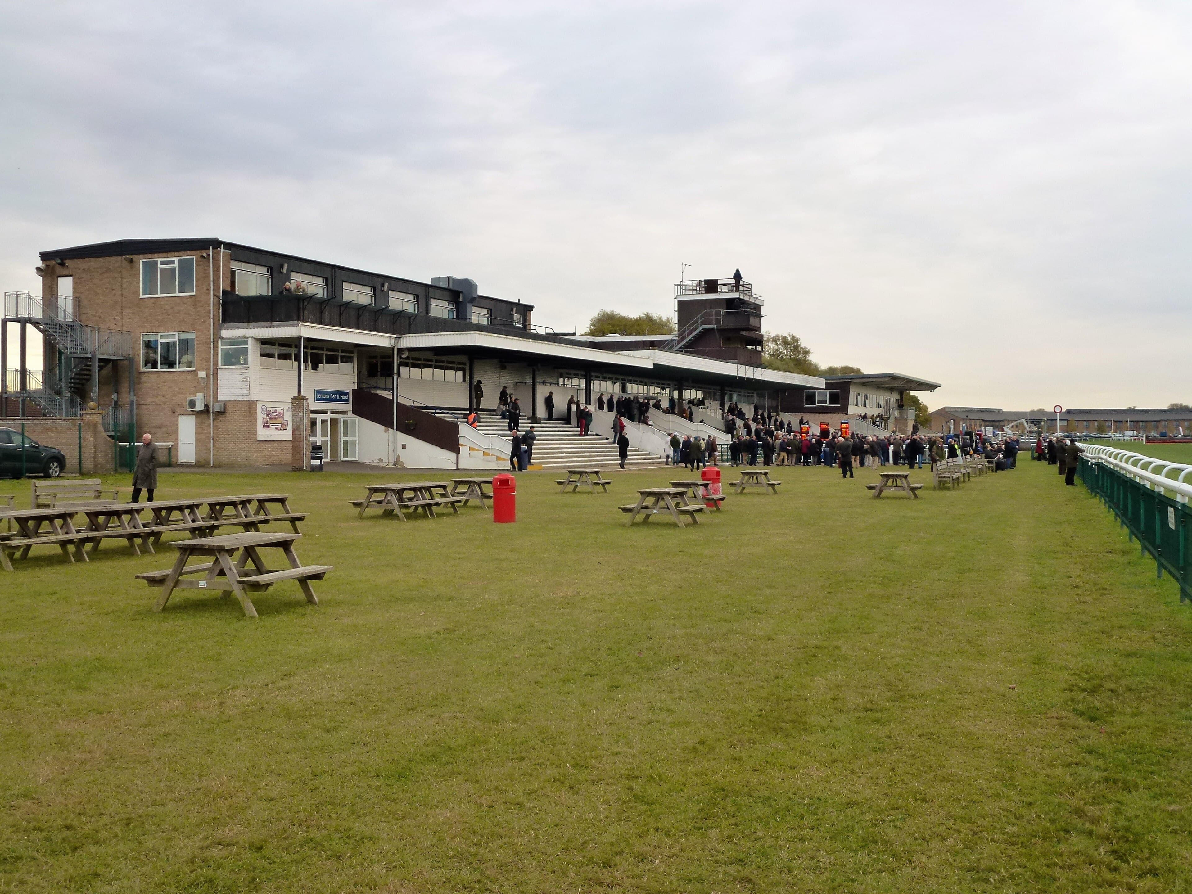 The long low grandstand at Huntingdon seen across the picnic tables on the lawn, racegoers gathered at the steps