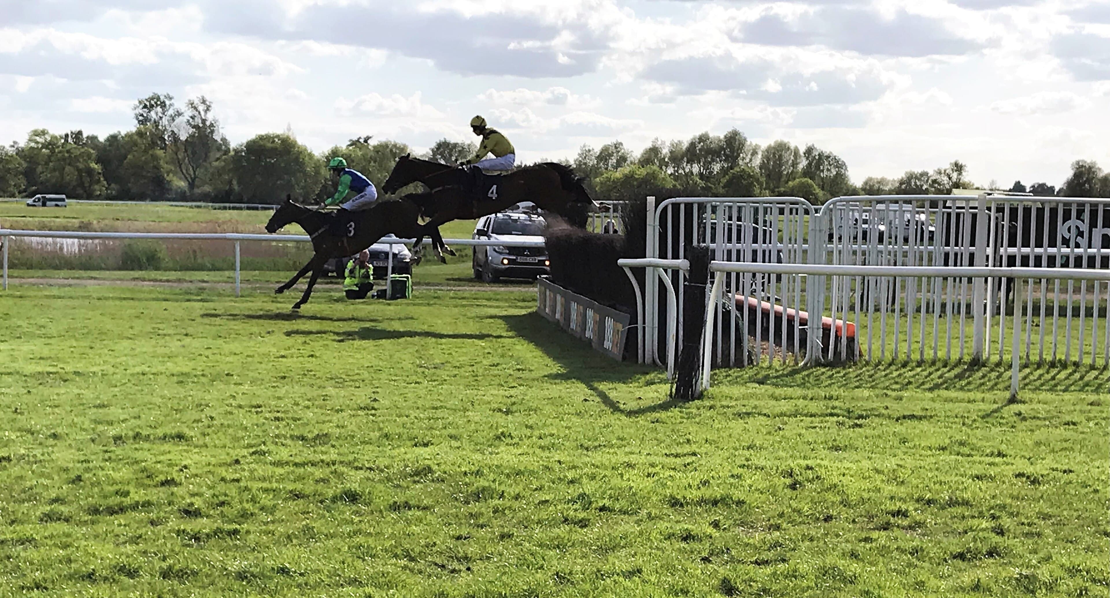 Two horses jumping the open ditch at Huntingdon side on, the birch fence and its guard rail running across the frame with the flat green course behind