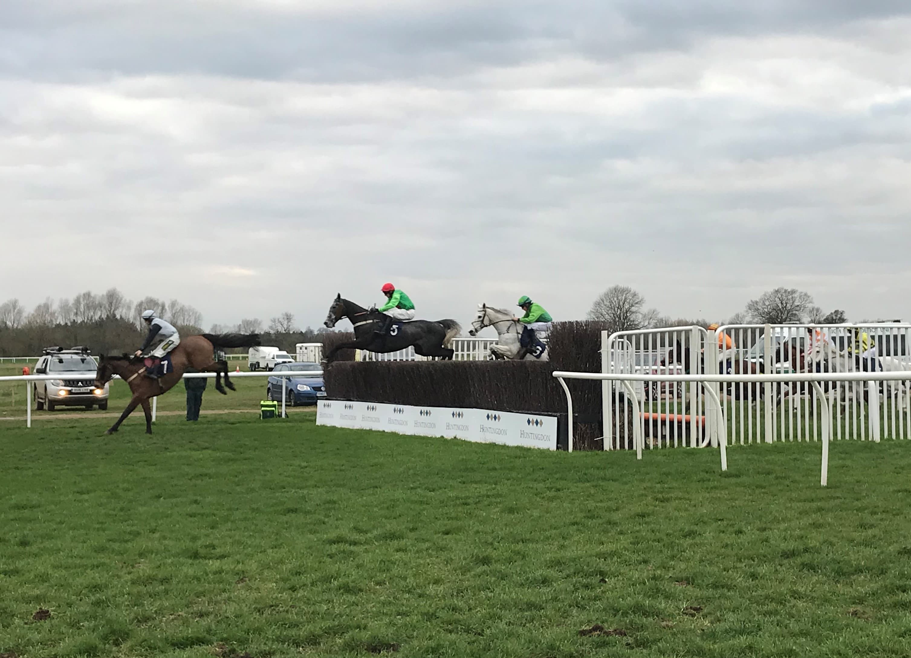 Two horses jumping the open ditch at Huntingdon while a third lands on the near side, the birch fence and its Huntingdon guard board running across the frame