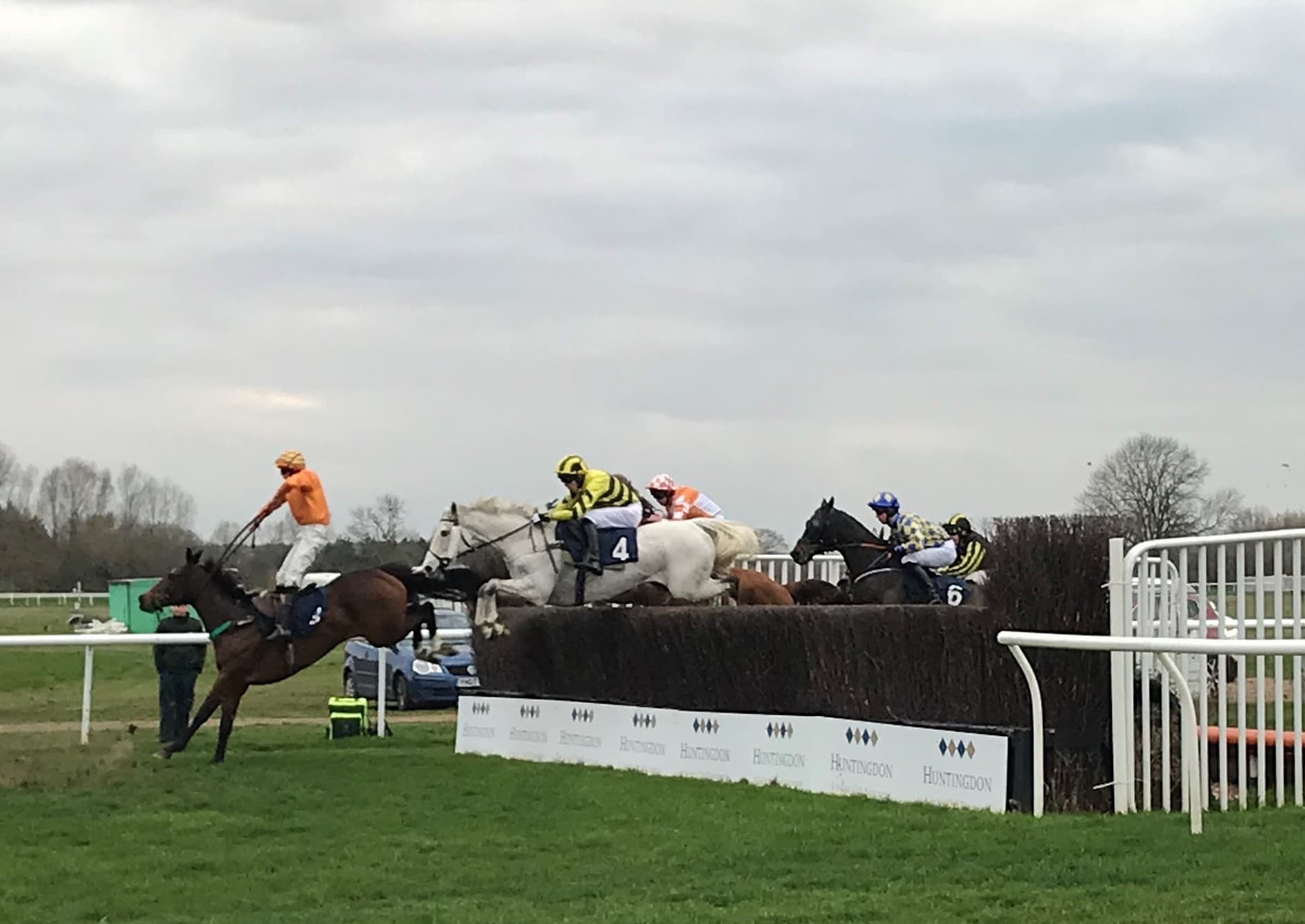Four horses and riders jumping a birch chase fence at Huntingdon, the take-off board carrying the racecourse name