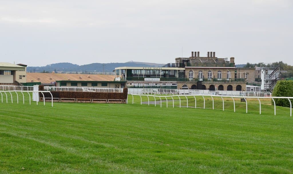A birch steeplechase fence in front of the stands at Kelso, with the Borders hills on the skyline.