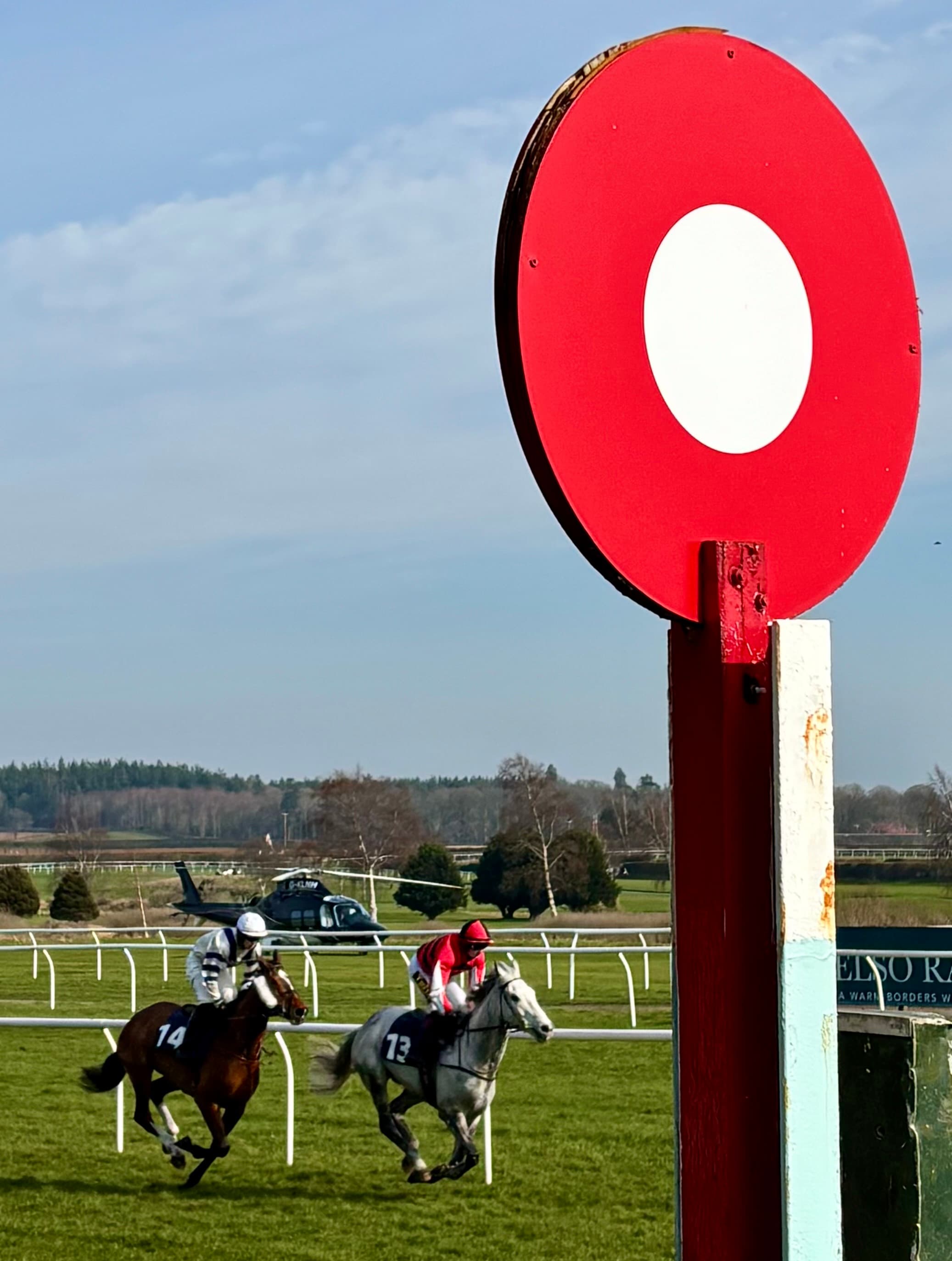 Two runners racing past the red-and-white winning post disc at Kelso on a clear spring afternoon.