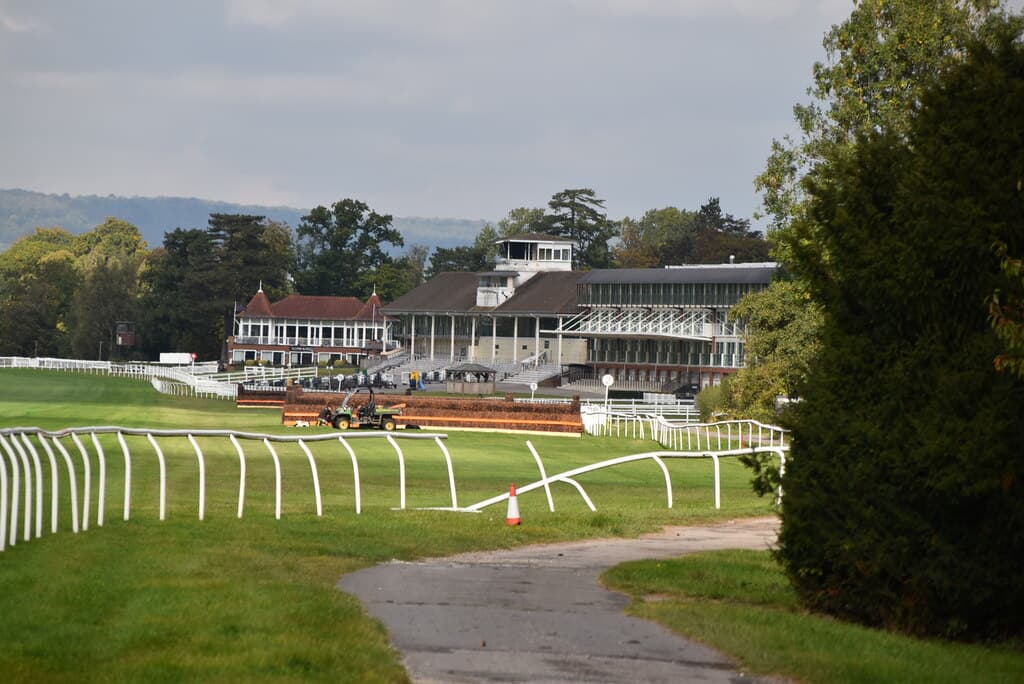 The grandstand at Lingfield Park seen from across the course, with a birch chase fence and a maintenance vehicle in front of it and the North Downs behind.