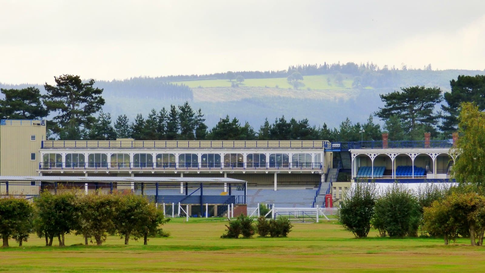 The glass-fronted grandstand and the older ironwork stand at Ludlow Racecourse, with the wooded Shropshire hills rising behind them.