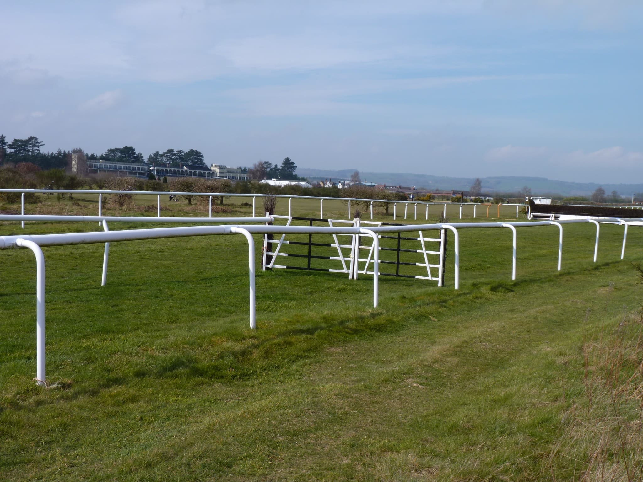 White running rails and a rail gate on a bend at Ludlow Racecourse, with a chase fence to the right and the grandstands in the distance.