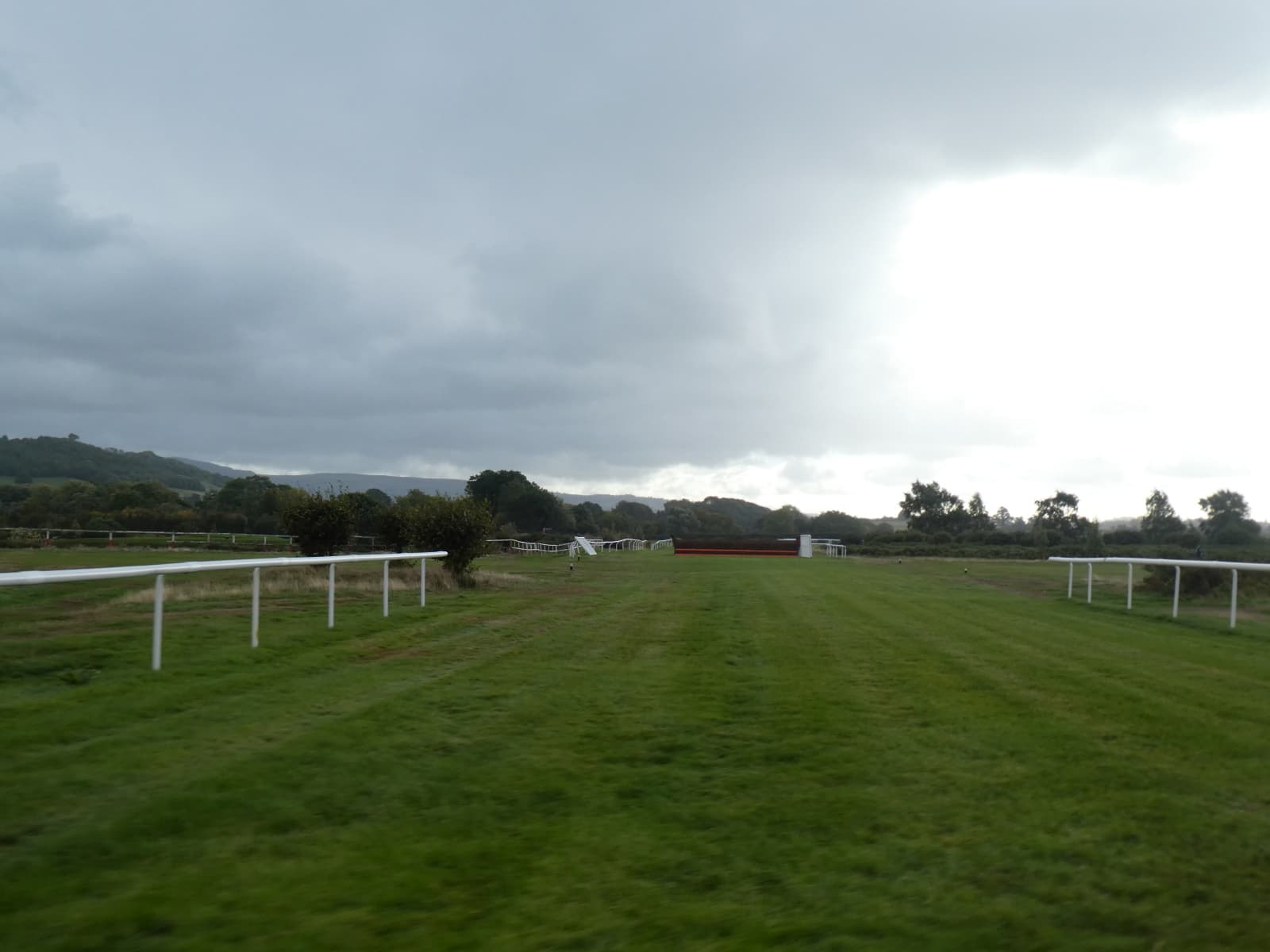 A birch chase fence standing across the track at Ludlow Racecourse under a heavy grey sky, with the running rails leading up to it.