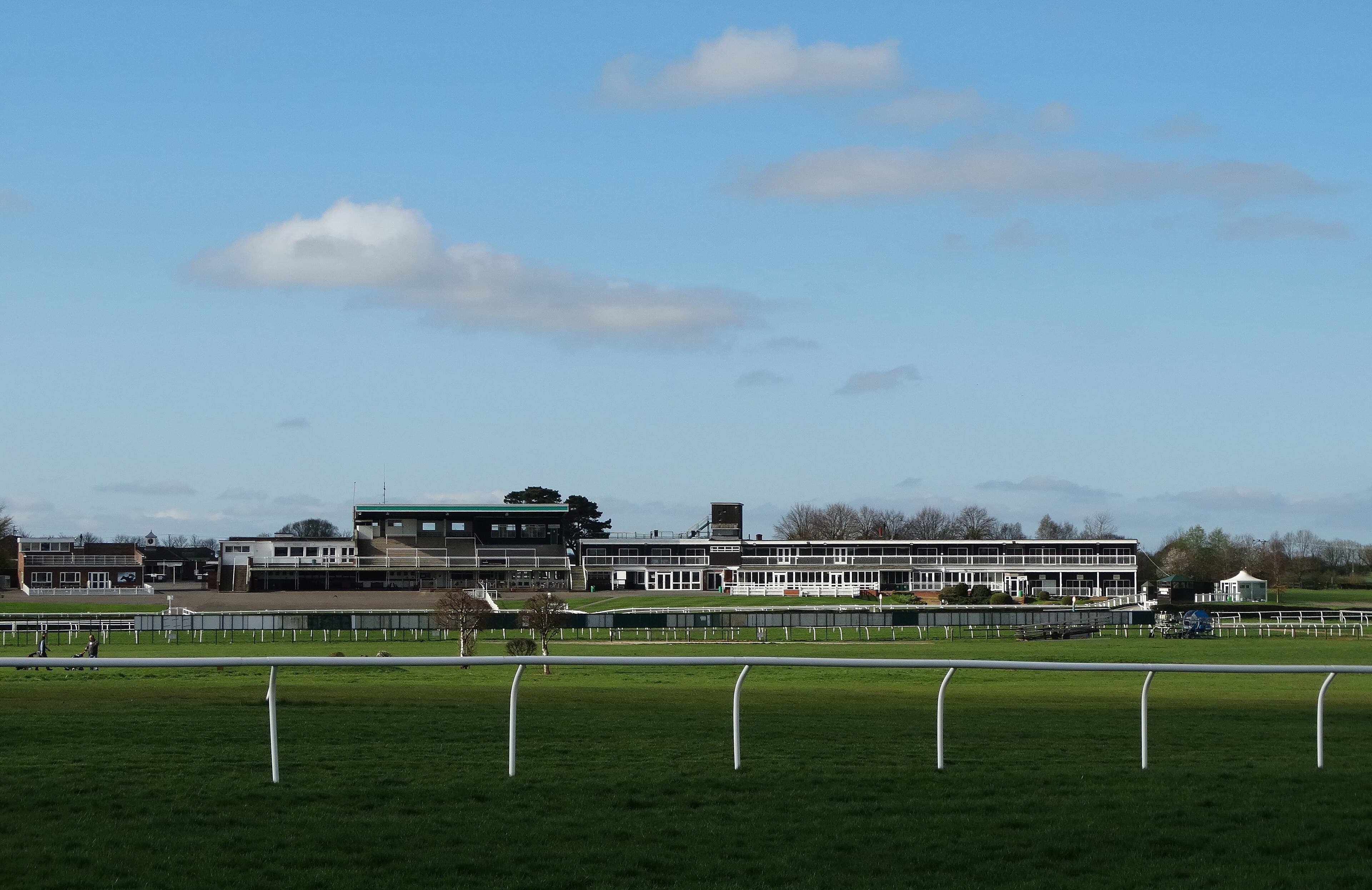 The stands at Market Rasen racecourse seen across the turf from the far side of the track, with the white running rail in the foreground under a blue sky