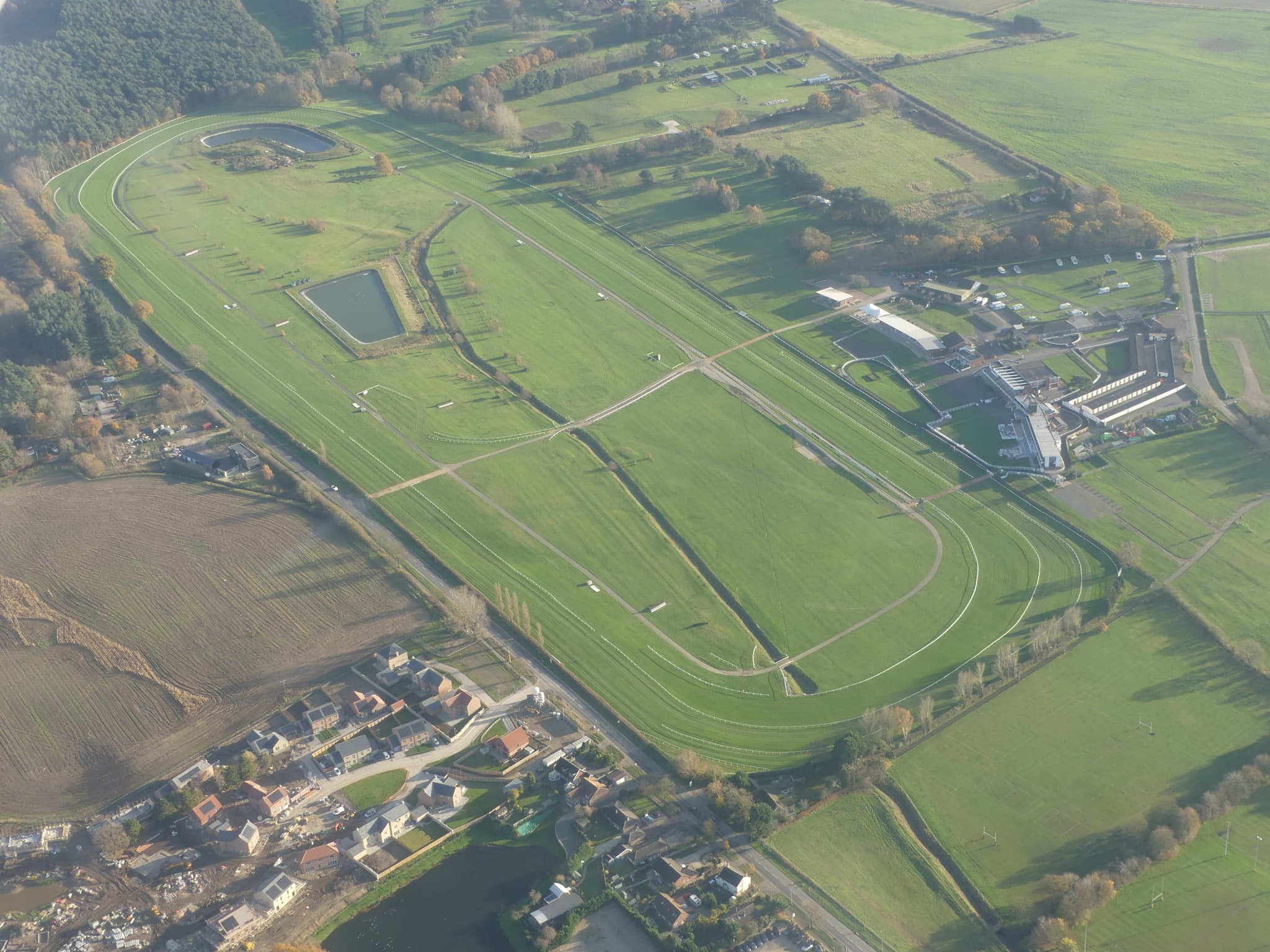 Aerial view of Market Rasen racecourse in autumn, showing the full oval of the jumps track with its fences and hurdles, the two lakes inside the course and the grandstand buildings on the far side