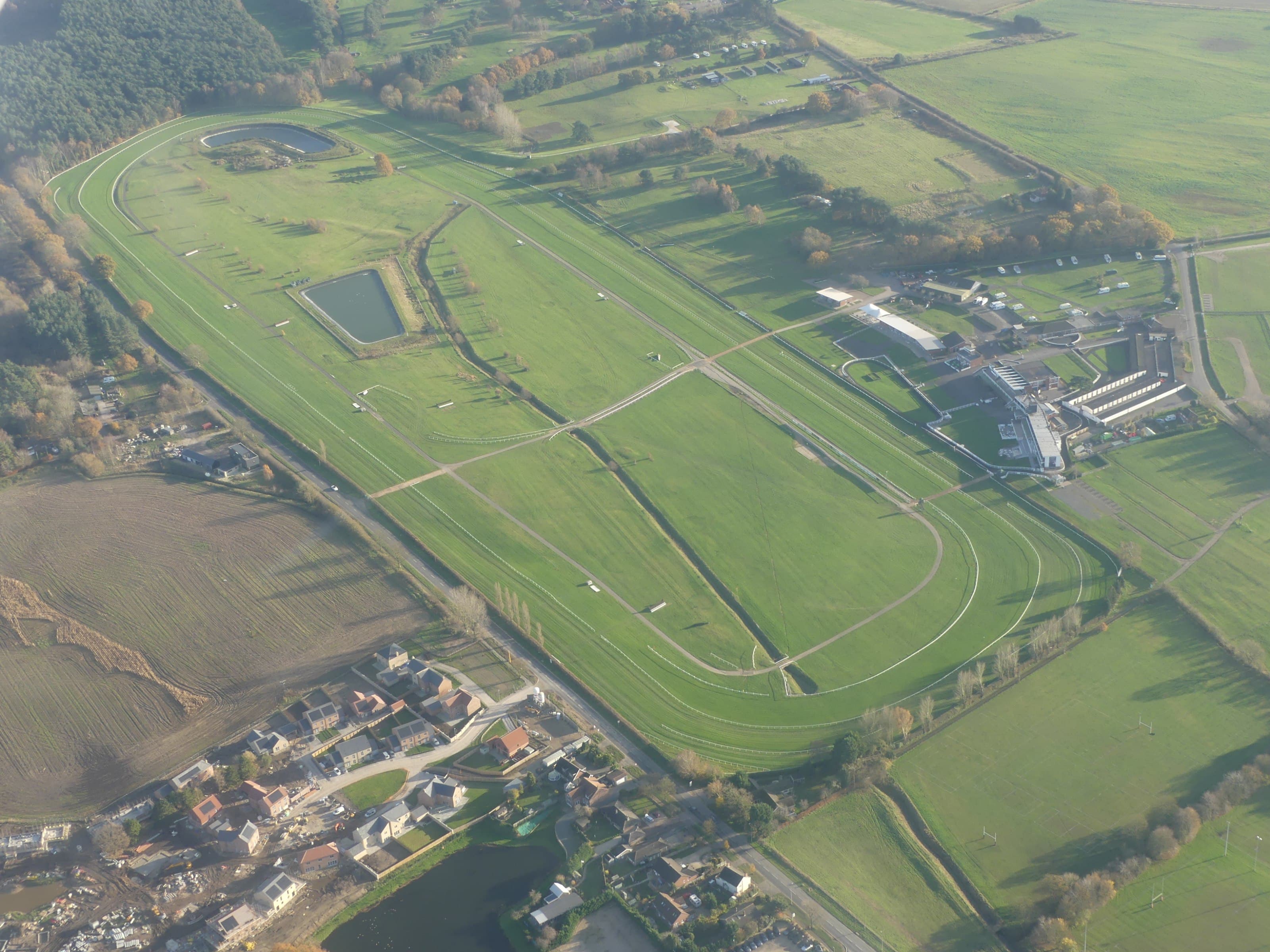 Aerial view of Market Rasen racecourse in autumn, showing the full oval of the jumps track with its fences and hurdles, the two lakes inside the course and the grandstand buildings on the far side