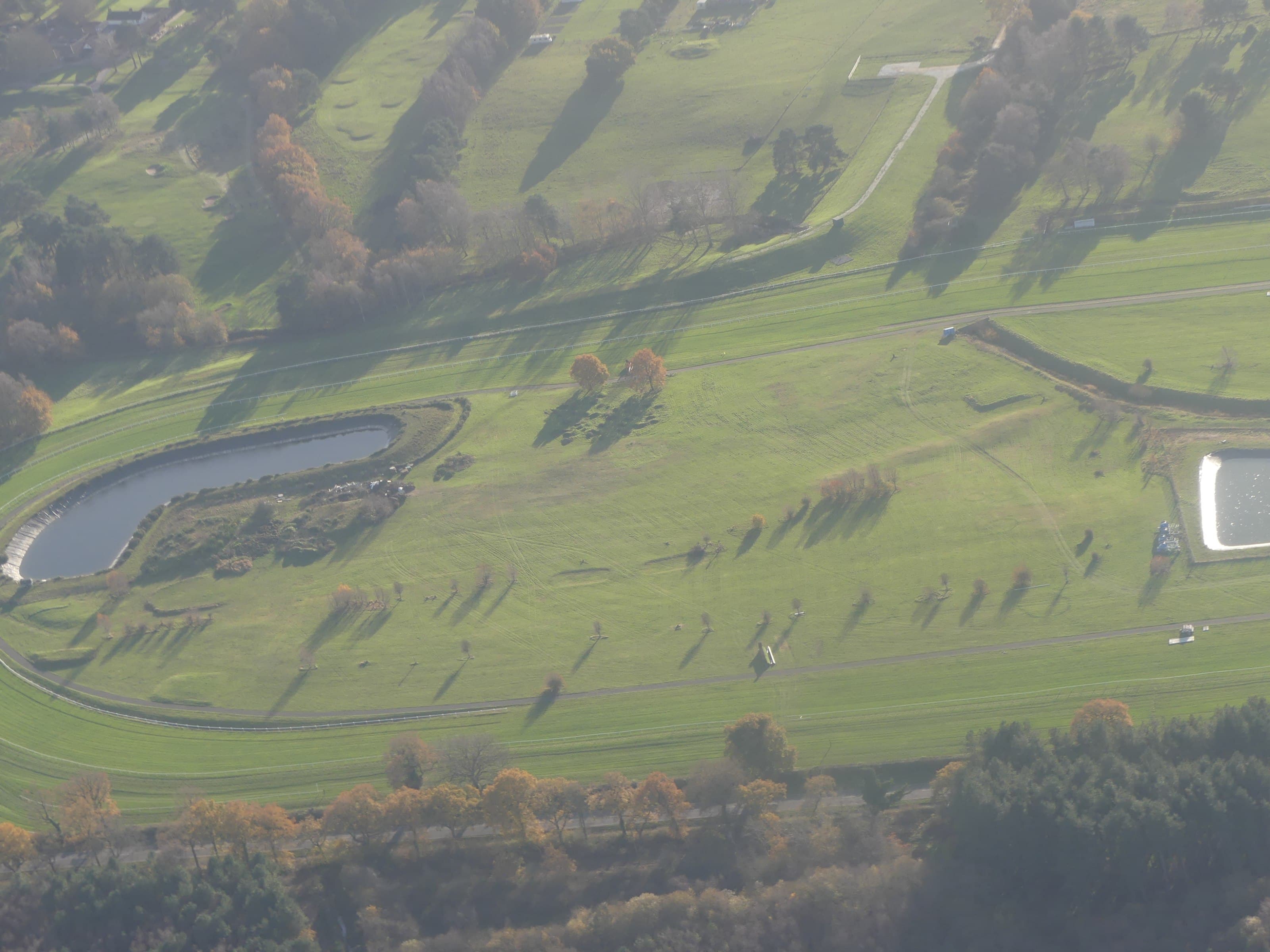 Aerial view of the far bend at Market Rasen racecourse in autumn, with the lake inside the running rail and the wooded ground beyond