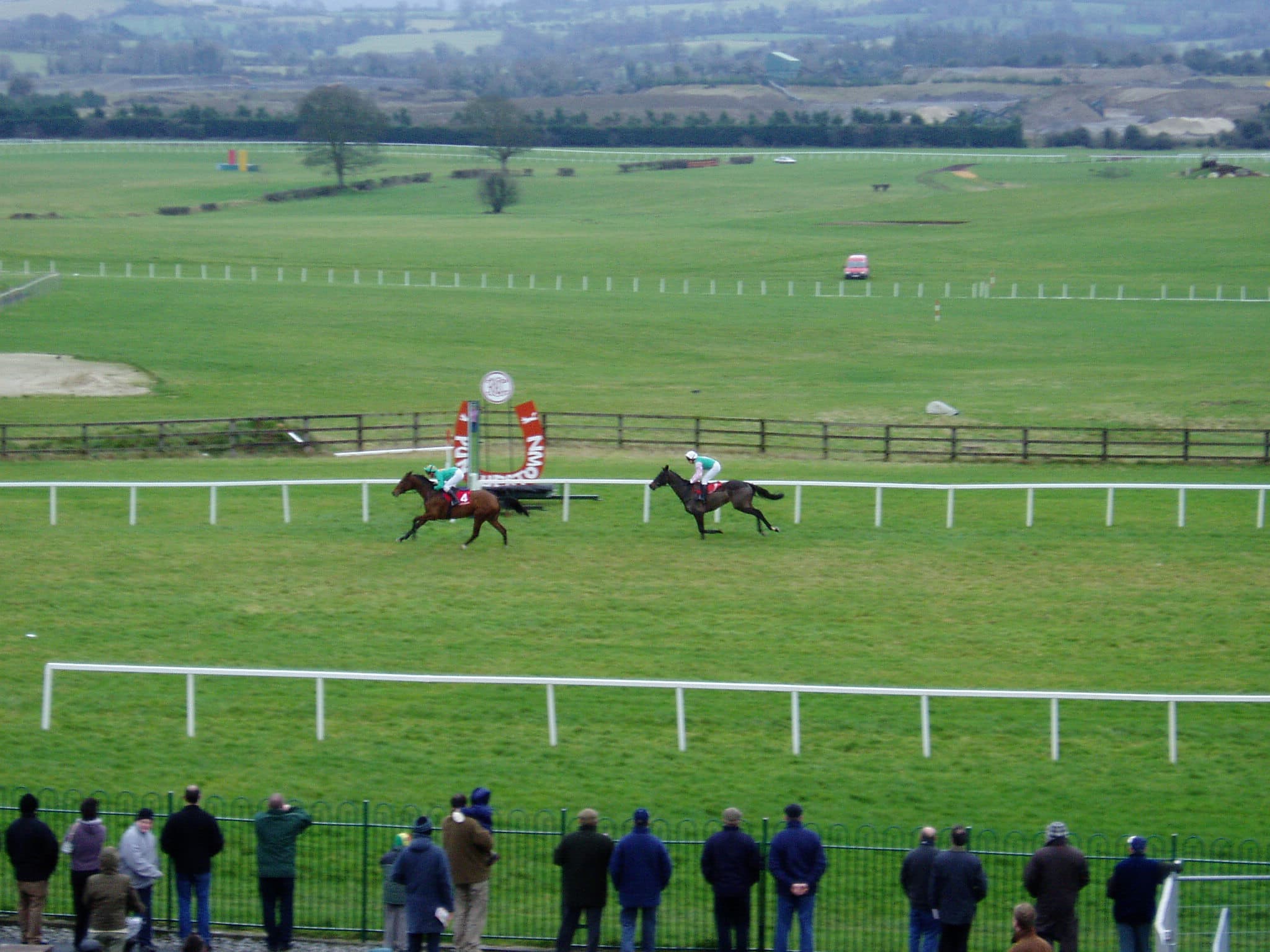 Two runners race away down the far side at Naas, spectators lined along the rail in the foreground and the Kildare country beyond.