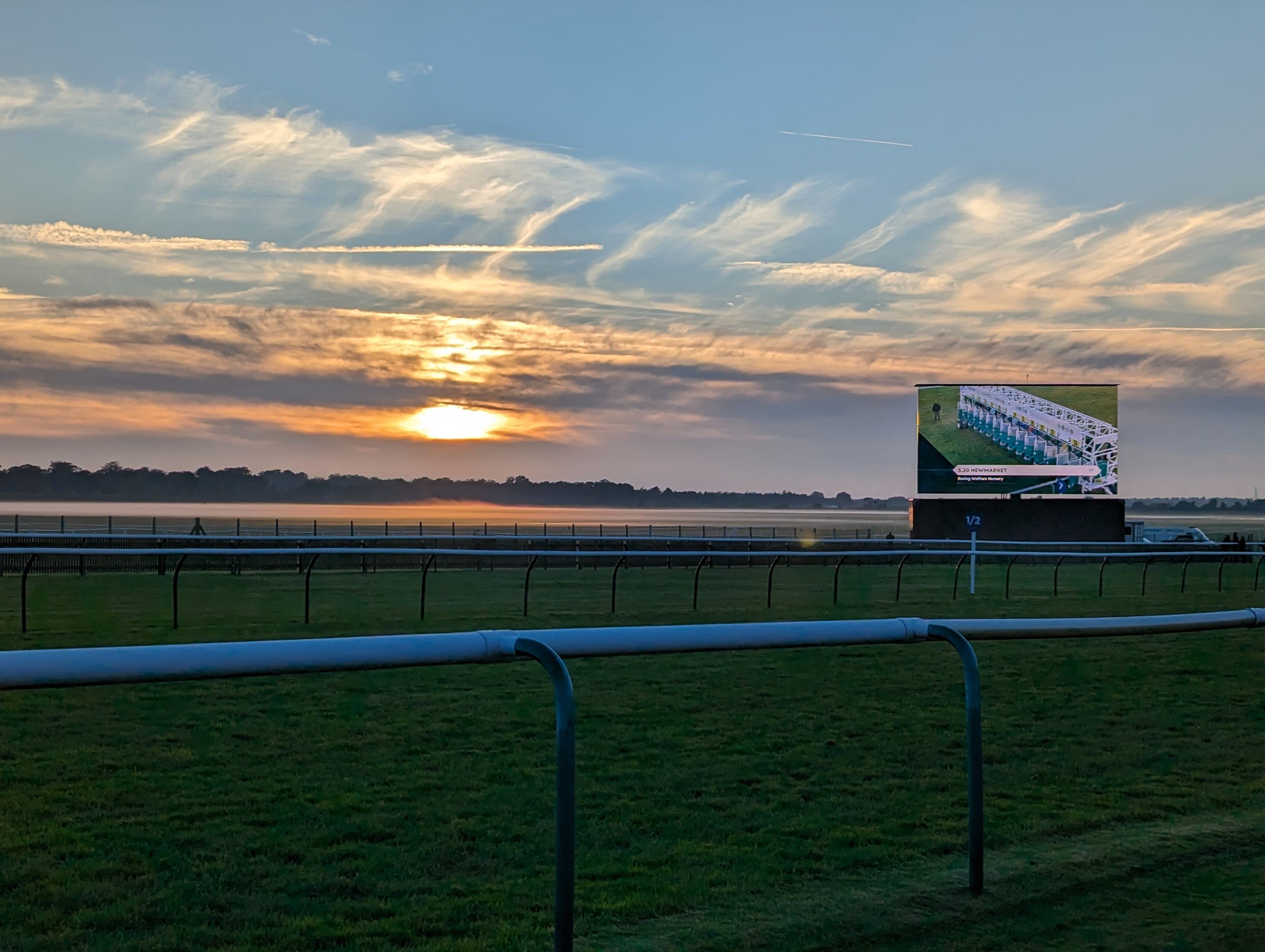Sunrise over the Rowley Mile at Newmarket, mist lying along the far rail and the big screen lit on the right.