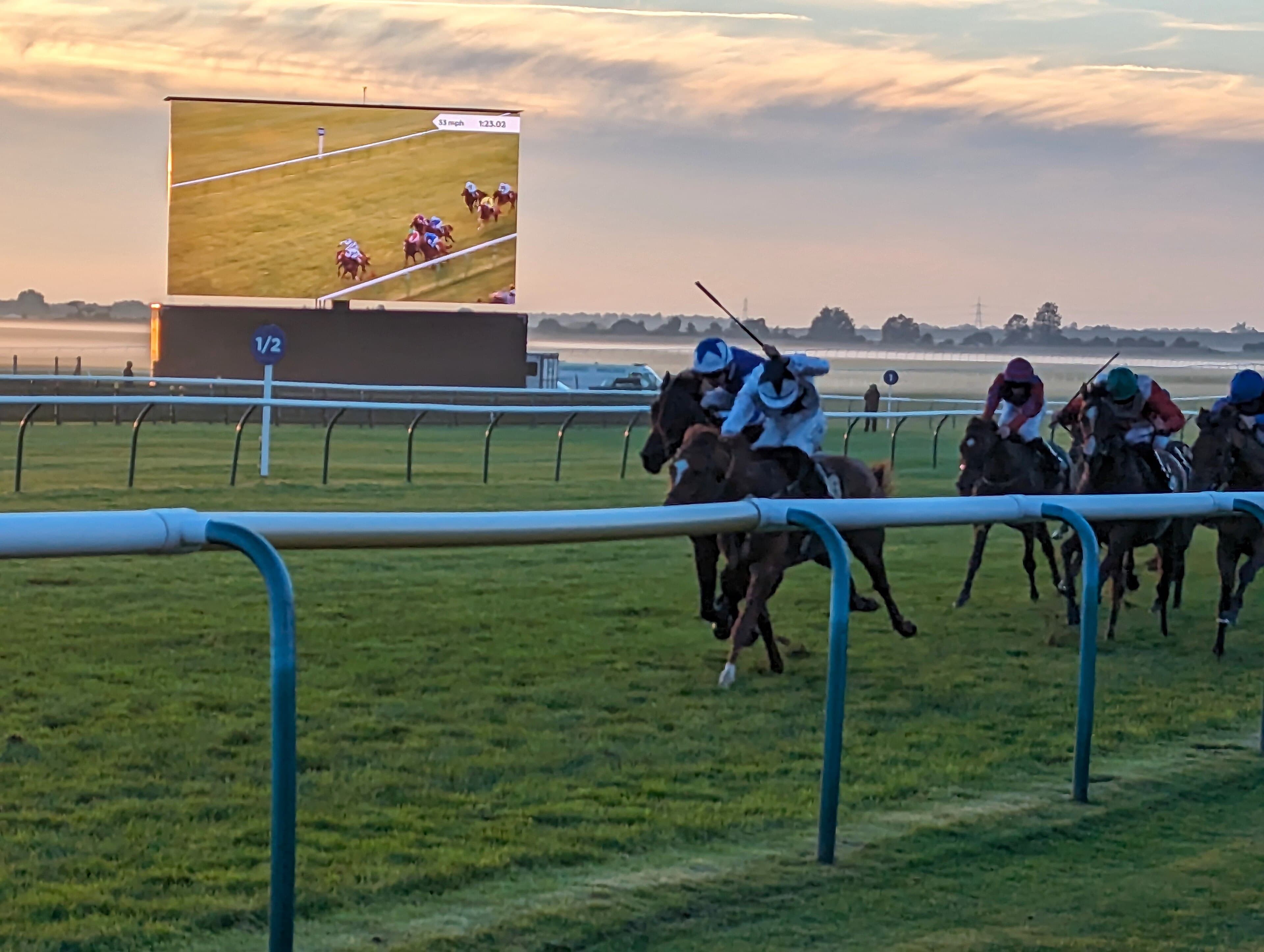Runners inside the last half furlong at Newmarket at sunrise, the big screen showing the race behind them.