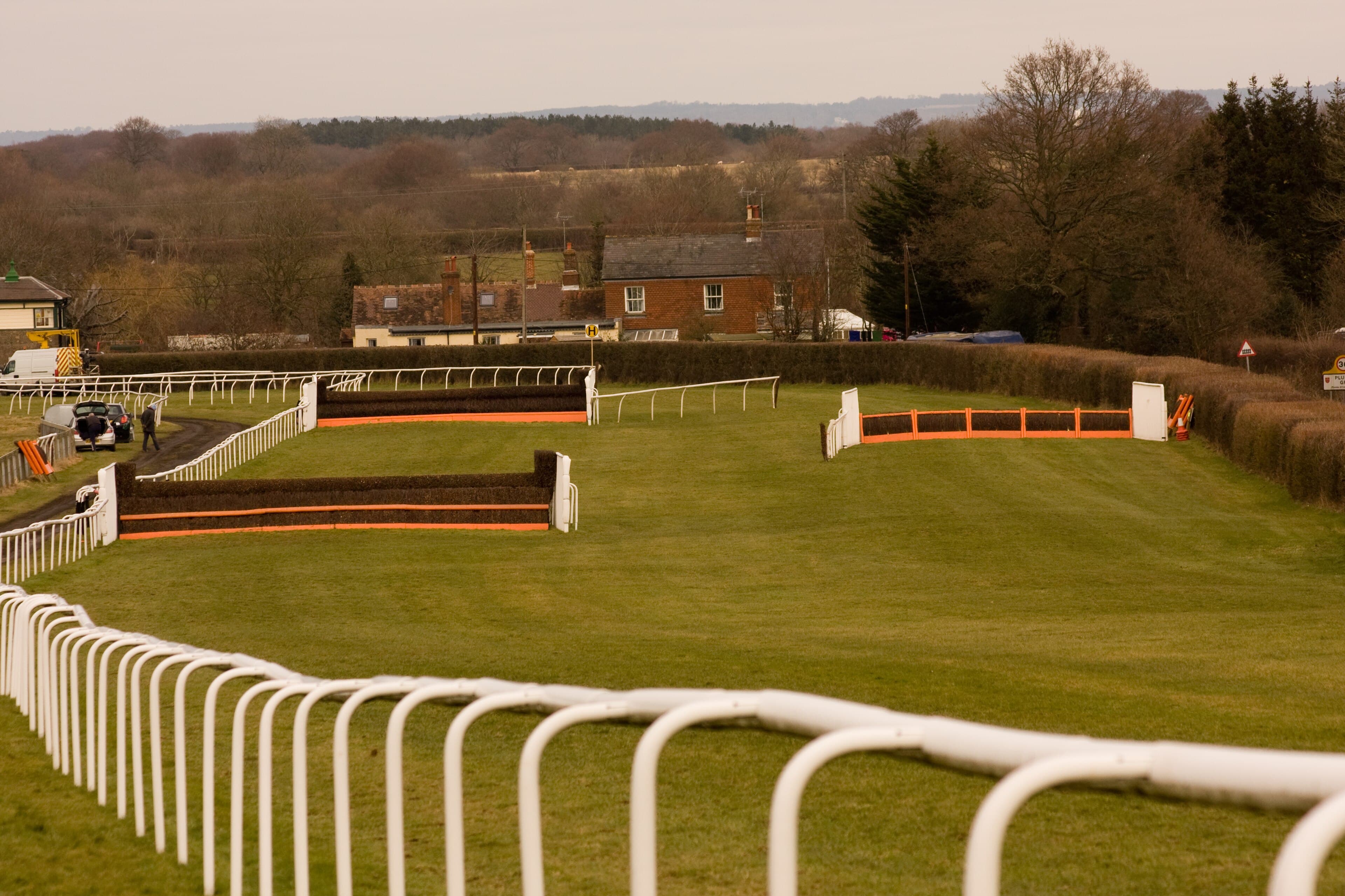 A hurdle and a chase fence in the back straight at Plumpton, cottages and bare trees on the boundary beyond.