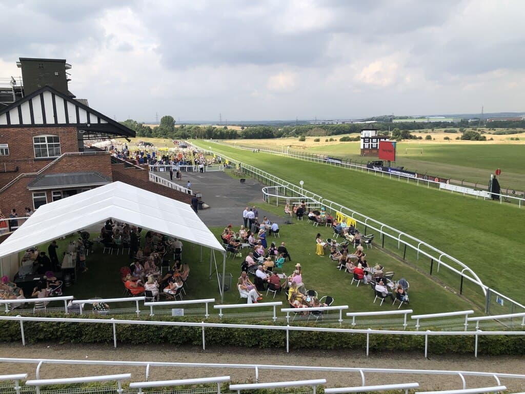 Racegoers at tables under a marquee beside the rail at Pontefract, the course and the big screen beyond.