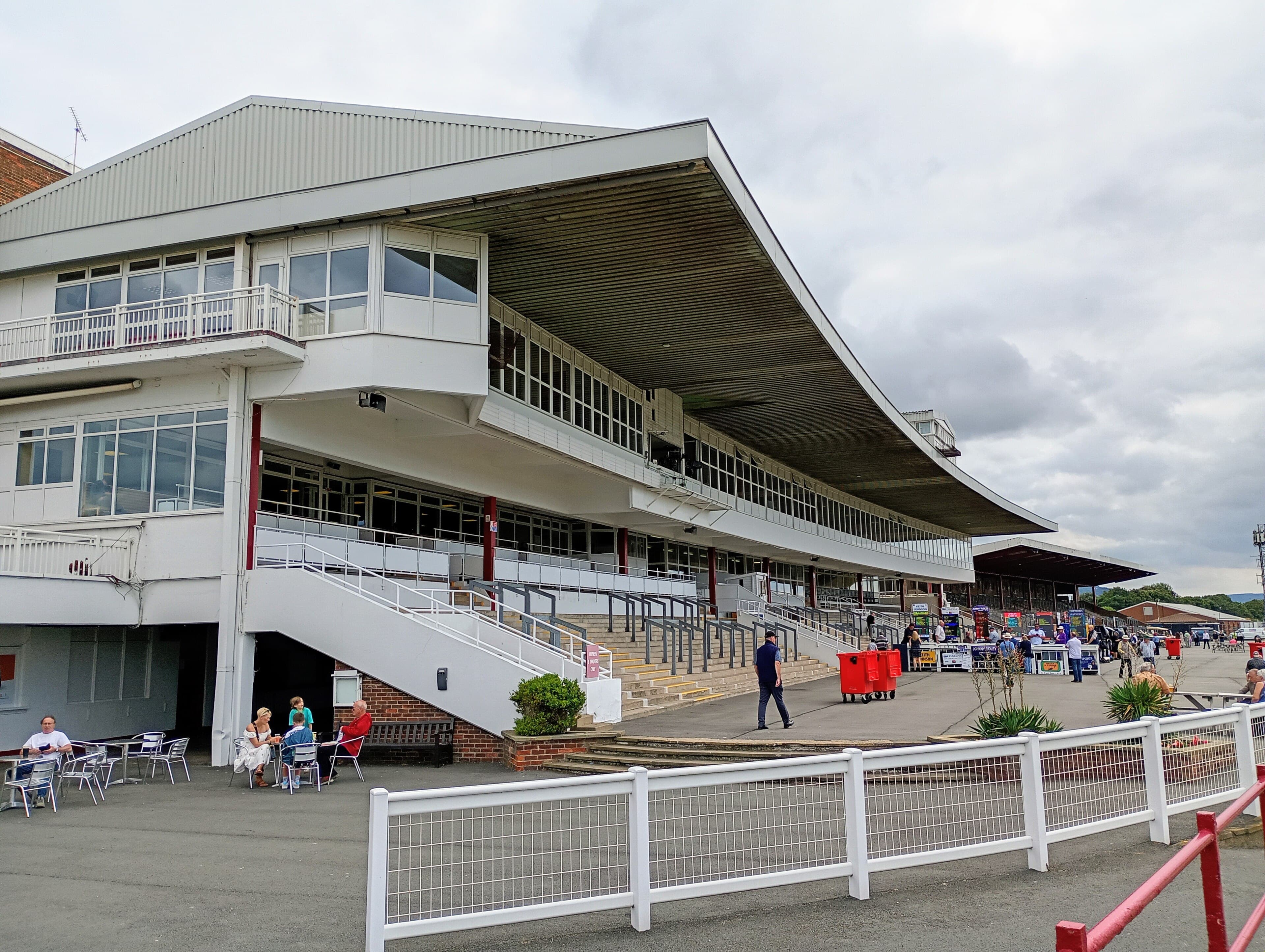 The cantilevered roof of the Redcar grandstand from the north, families at tables on the apron below.