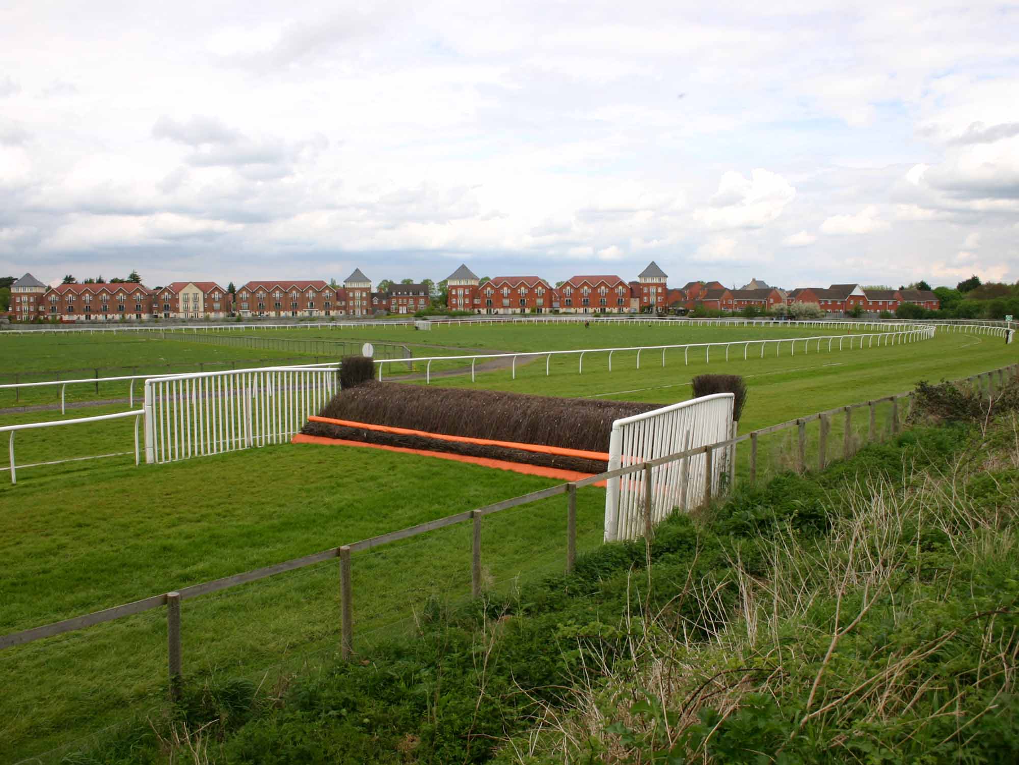 A birch chase fence with its orange take-off board at Stratford, the housing along Shottery beyond the far rail.
