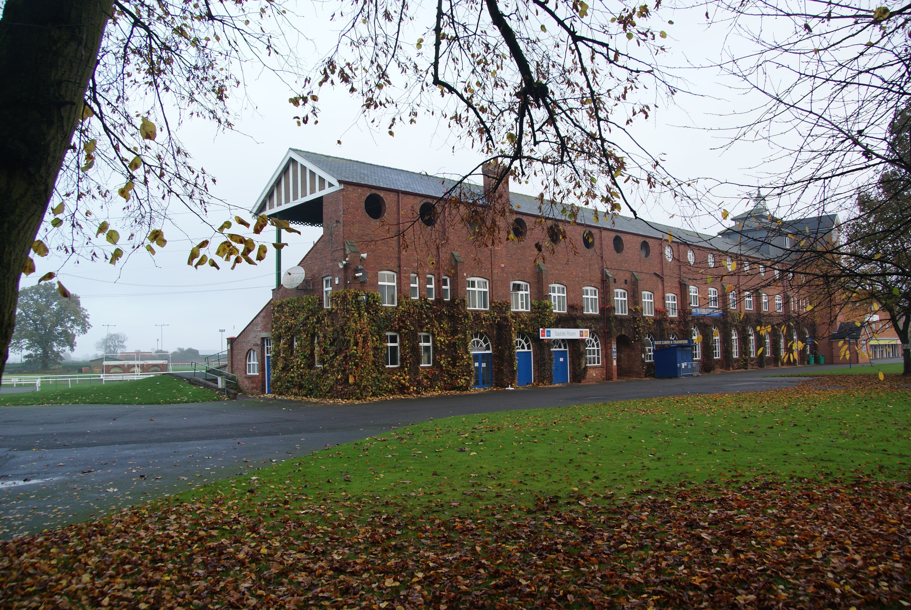 The ivy-covered red-brick range at Thirsk Racecourse under autumn trees, fallen leaves across the apron.