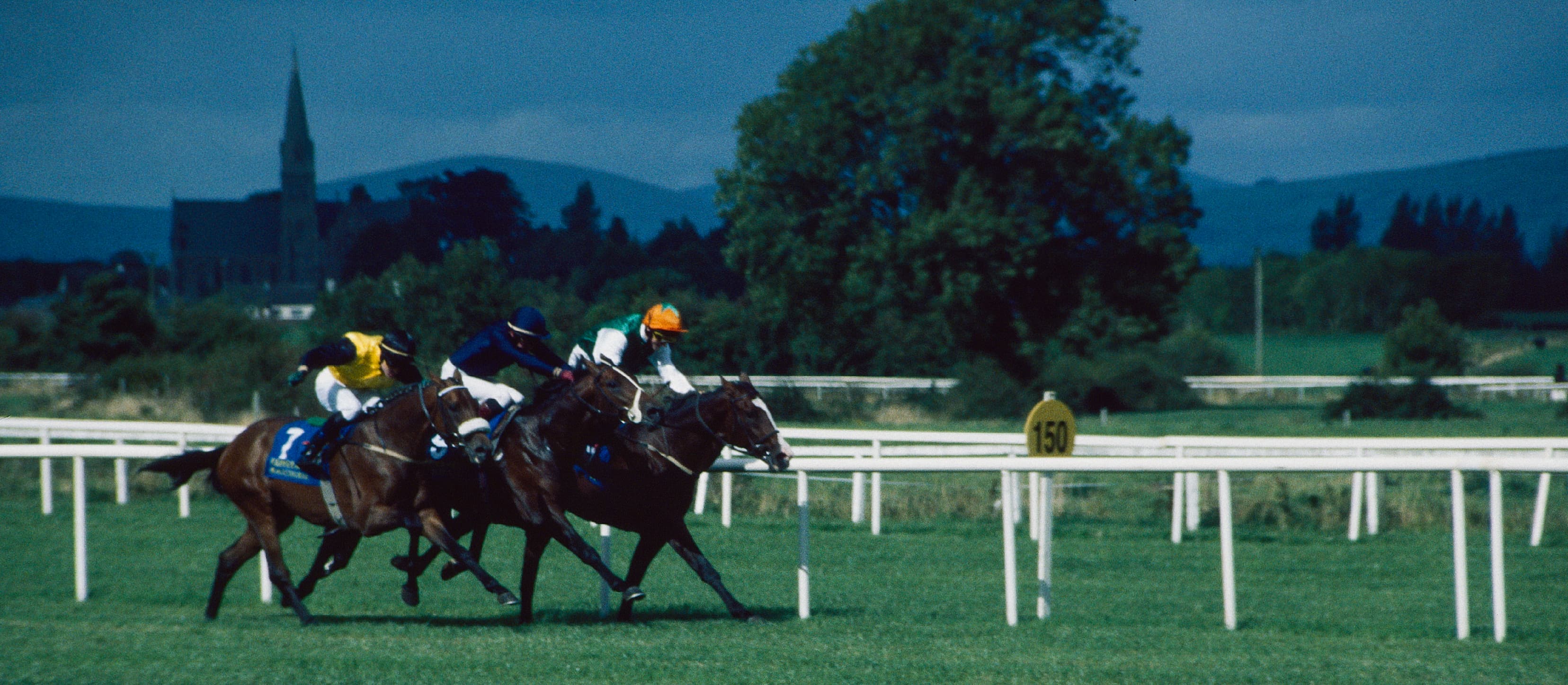 Three runners race up the straight at Tipperary past the 150 yards marker, a church spire and the Galtee hills behind.