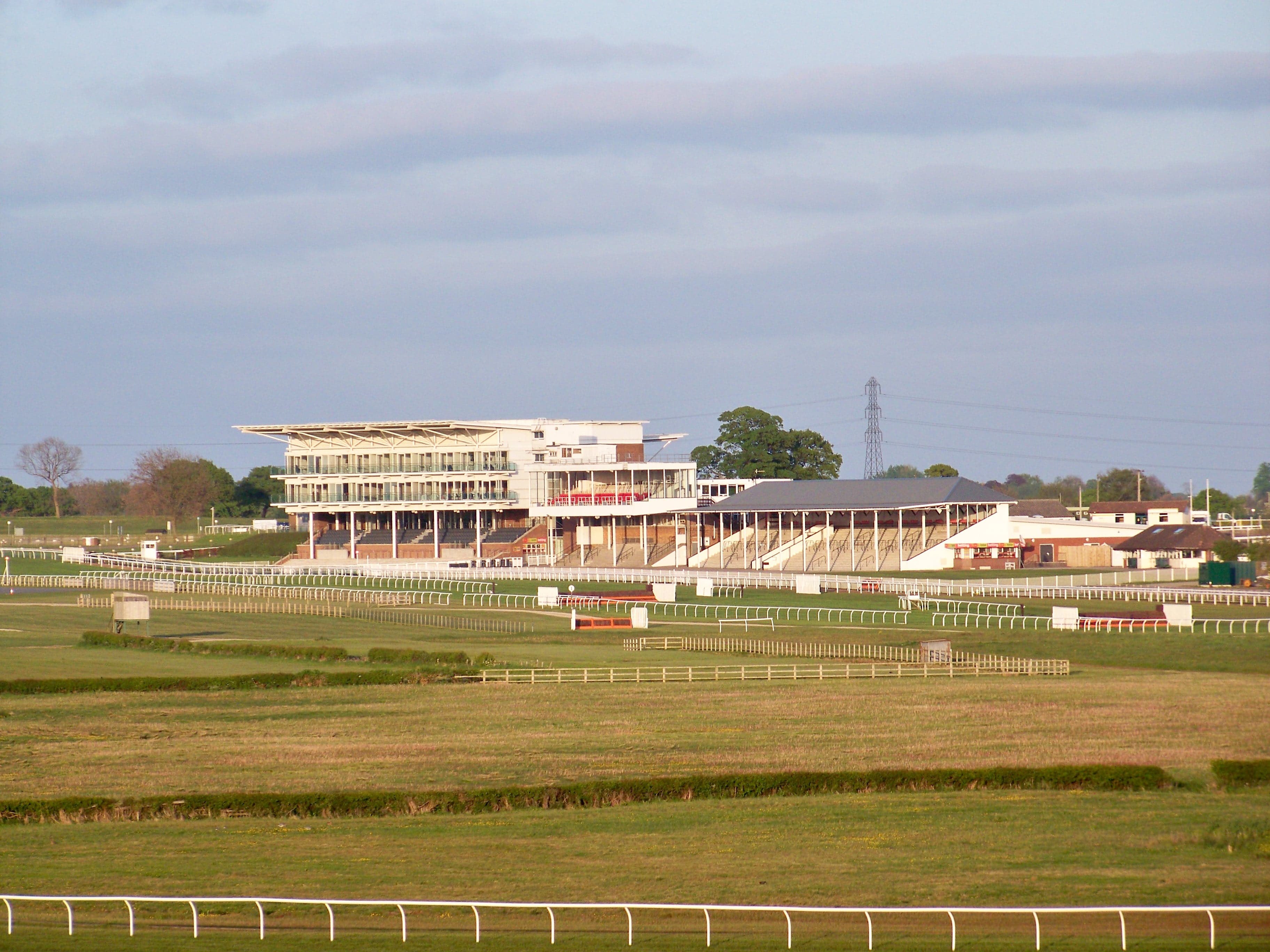 The stands at Wetherby racecourse in evening light, with the running rails and birch fences of the jumps course in front of them.