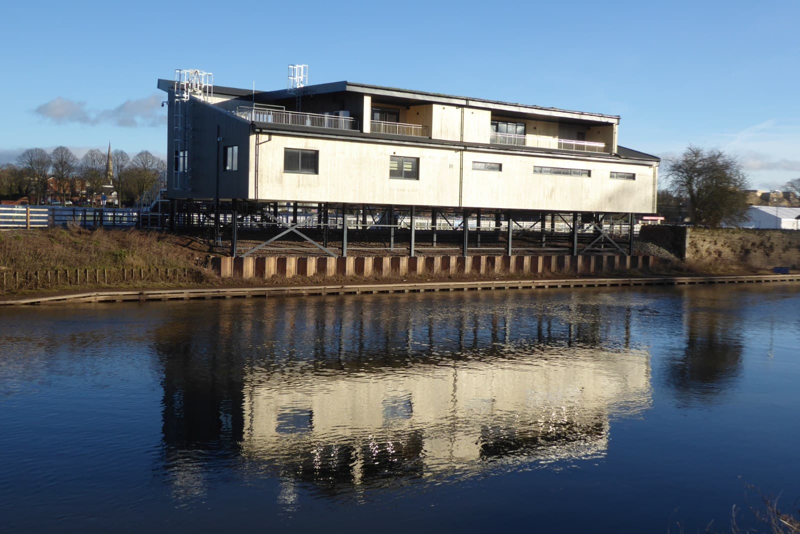 The weighing room and stand at Worcester racecourse raised on stilts above the bank of the River Severn, reflected in the water.